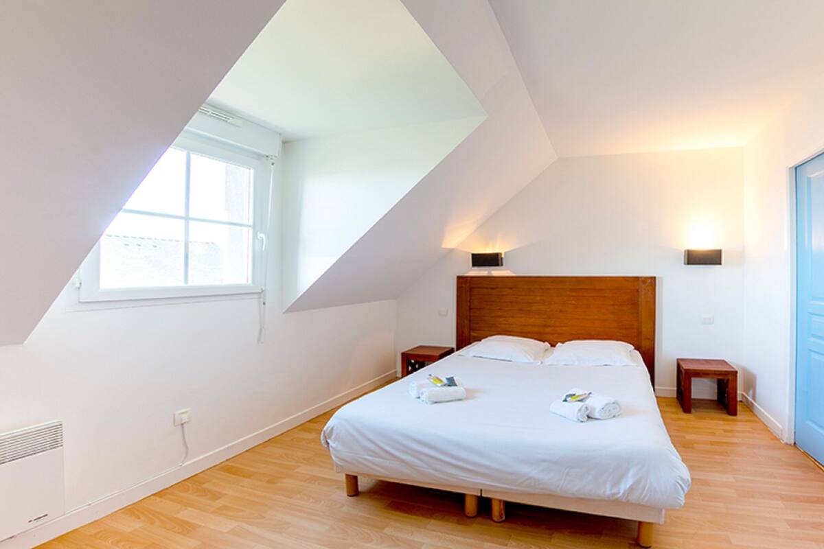 Un dormitorio con techo inclinado, cabecera de madera, cama grande con ropa de cama blanca, dos mesitas de noche y una ventana.