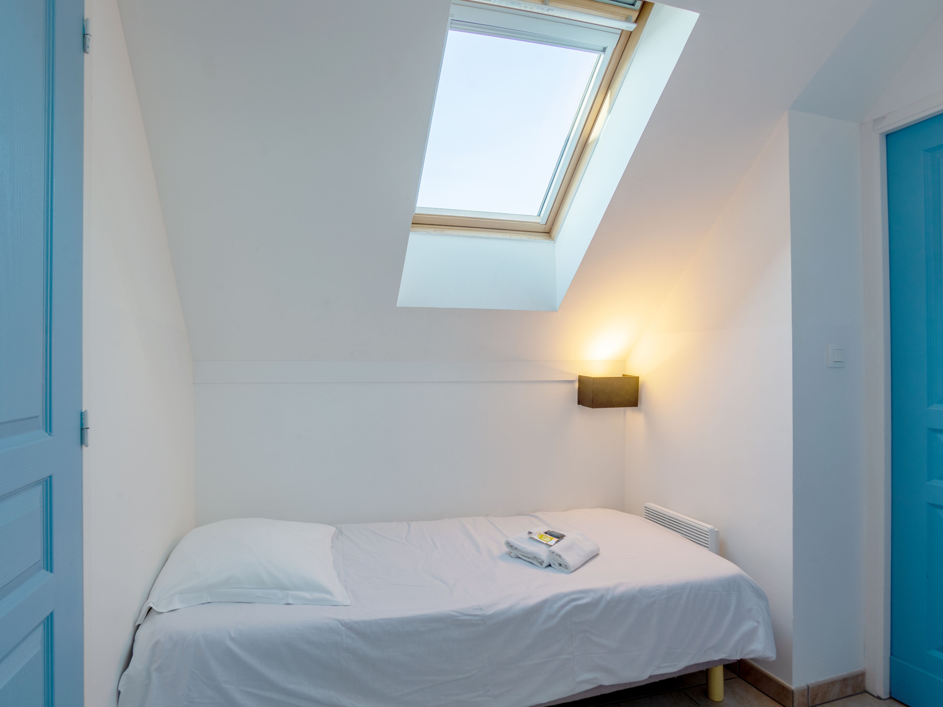 Ein kleines Zimmer mit einem Einzelbett, einem Dachfenster, einer Wandleuchte und einer Heizung.