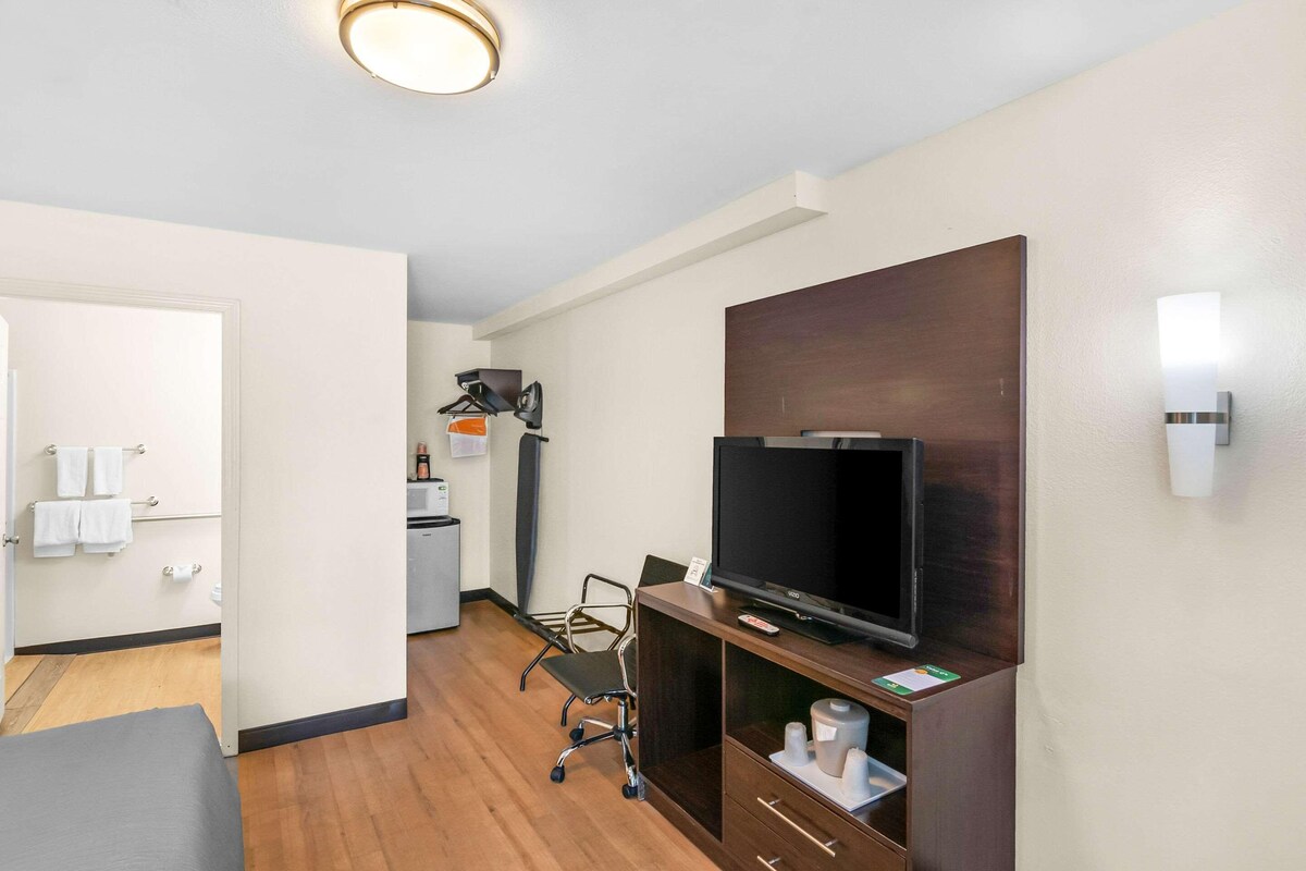 A hotel room with a flat-screen TV, a desk, a chair, a small kitchenette, and a bathroom visible through an open door.
