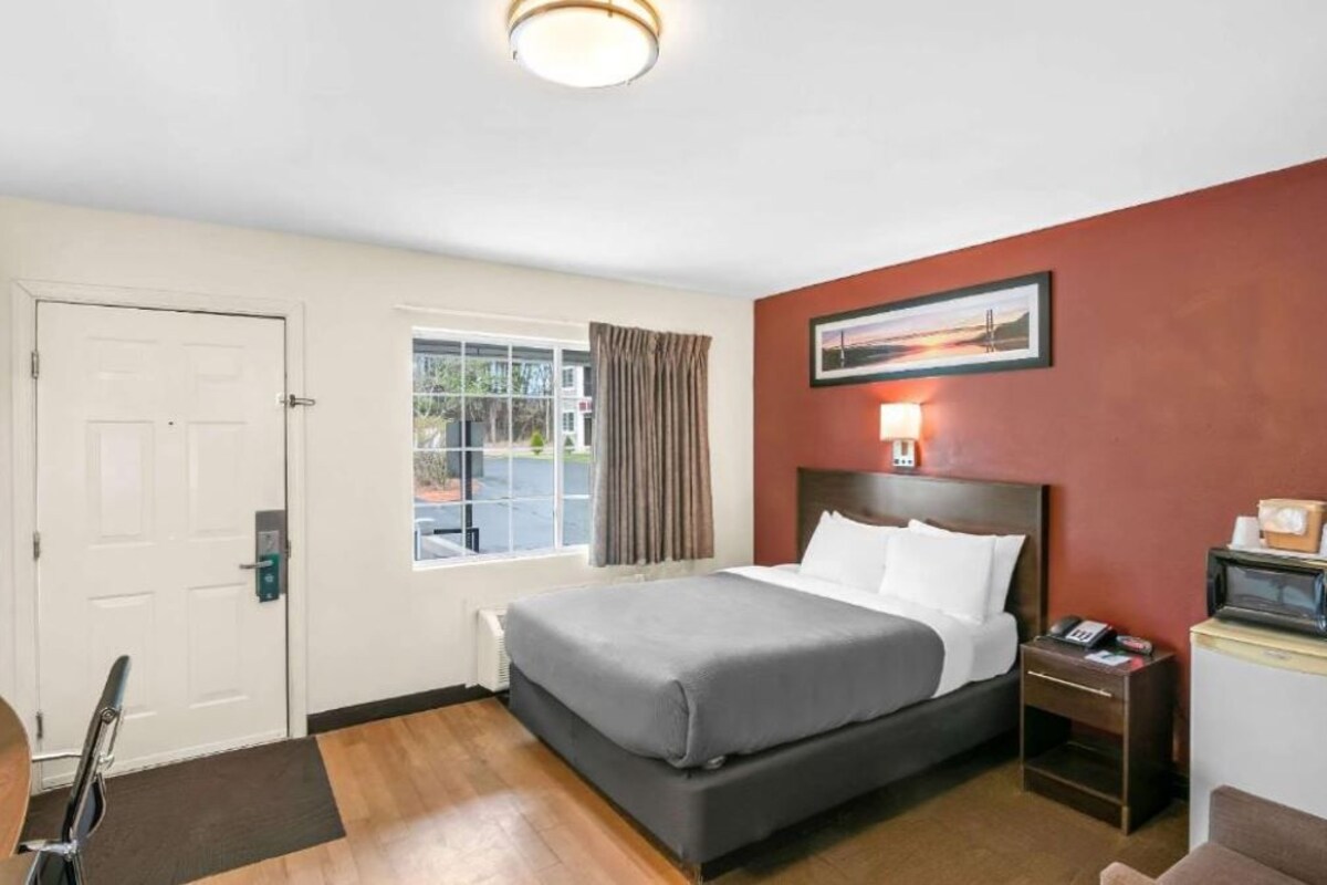 Una habitación de hotel con cama, escritorio, silla, ventana con cortinas, puerta y un cuadro en la pared.