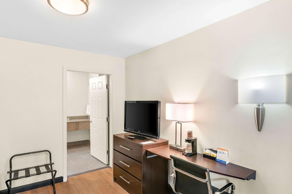 A hotel room with a desk, a television, a lamp, a chair, and a small table with a WiFi sign.