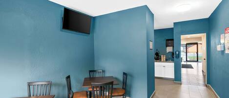 Un comedor con una mesa y sillas, un televisor montado en la pared y una cocina visible a través de un vano abierto.