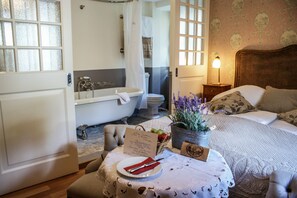 Un dormitorio con cama, mesita de noche con lámpara, baño con bañera y ventana con cortinas.