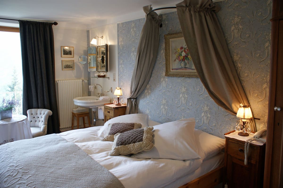 Un dormitorio con cama, mesitas de noche, sillón, ventana con cortinas, radiador, espejo y una pared decorada con un cuadro.