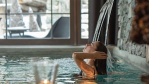 Une personne se détend dans un bain à remous avec une cascade en arrière-plan, un verre d’eau à portée de main et un mur de pierres en fond.