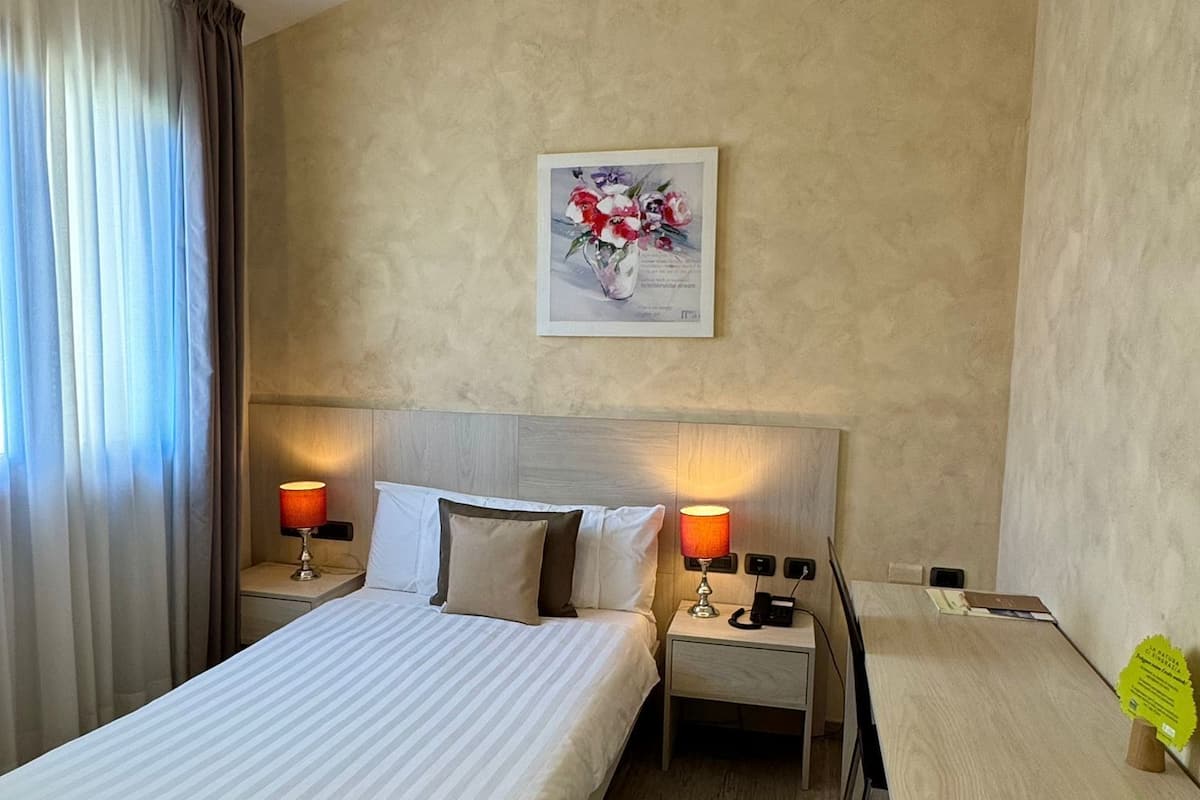 Habitación de hotel con una cama, dos mesitas de noche con lámparas, un cuadro en la pared y un escritorio con una silla.