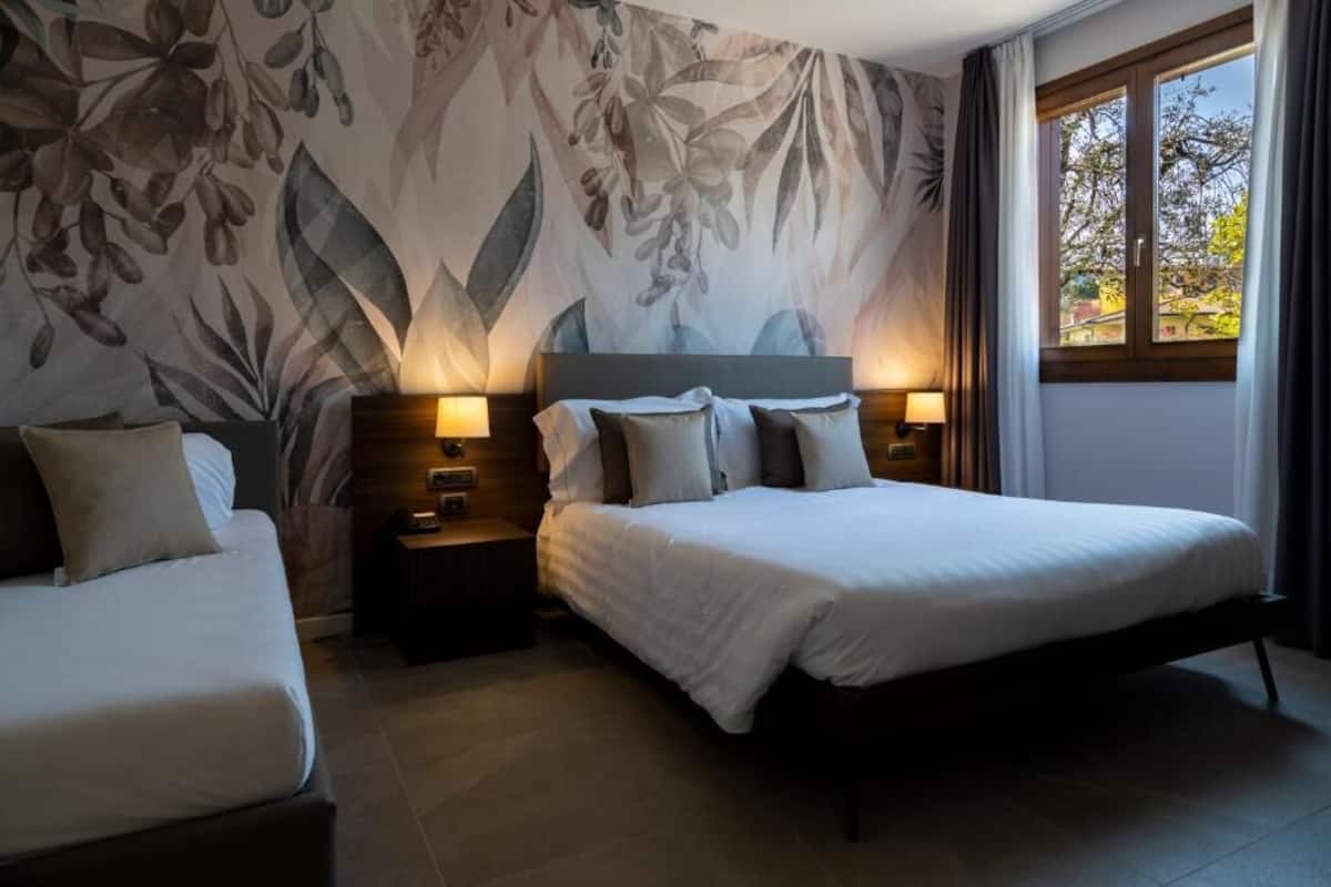 Habitación de hotel con una cama grande, dos mesitas de noche con lámparas, un mural floral en la pared y una ventana con cortinas.