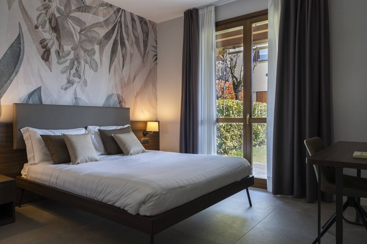 Habitación de hotel moderna con una cama grande, una mesita de noche, una mesa y vistas al exterior a través de un ventanal amplio.