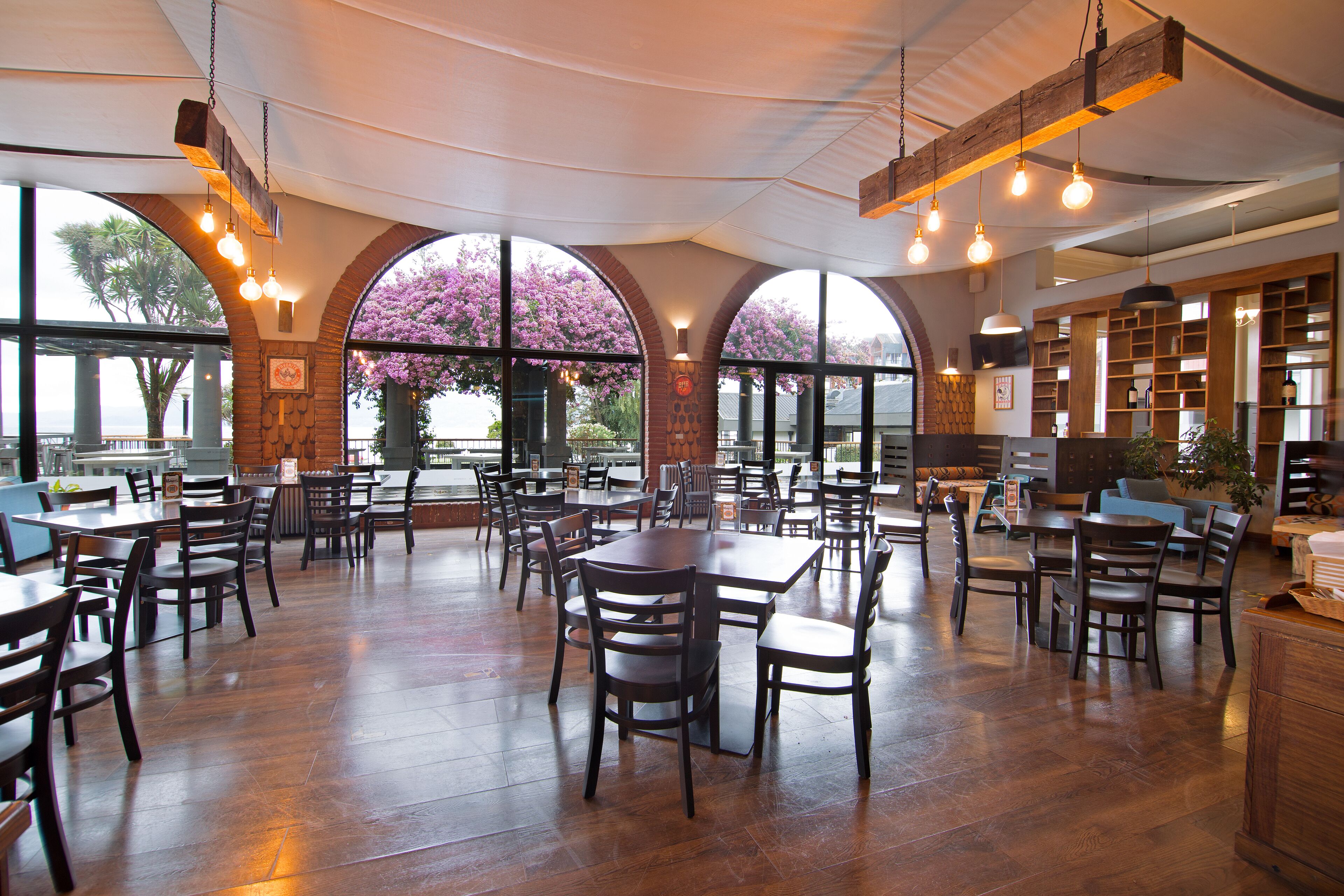 A spacious dining area with multiple tables and chairs, large windows with a view of trees, and hanging light fixtures.