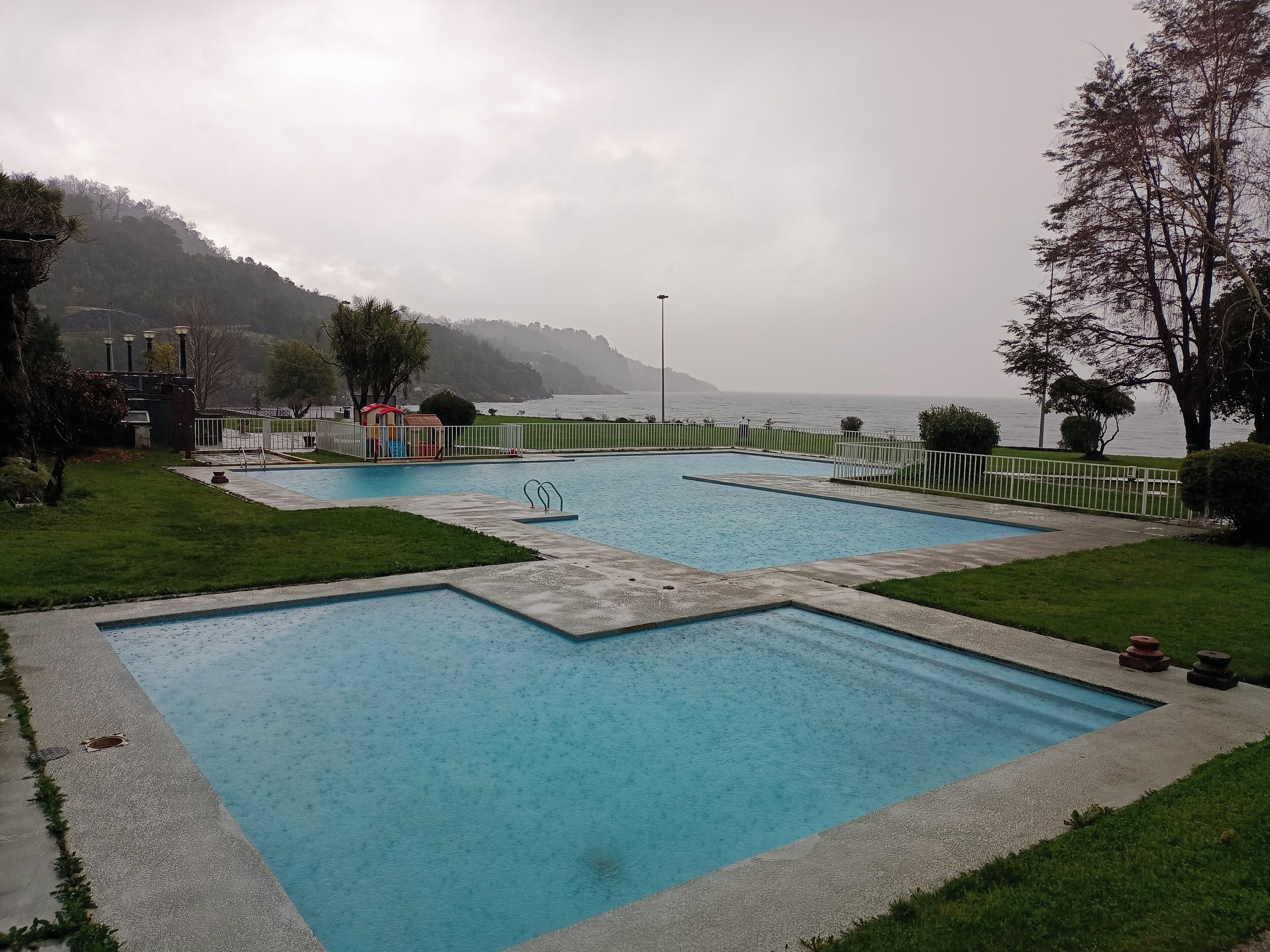 A rectangular pool with a surrounding deck, a small play area, and a view of the sea and hills in the background.