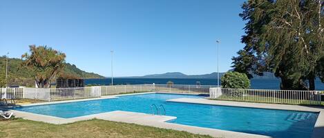 Una piscina rectangular con una cerca blanca, rodeada de césped y árboles, con vistas despejadas al mar y colinas al fondo.