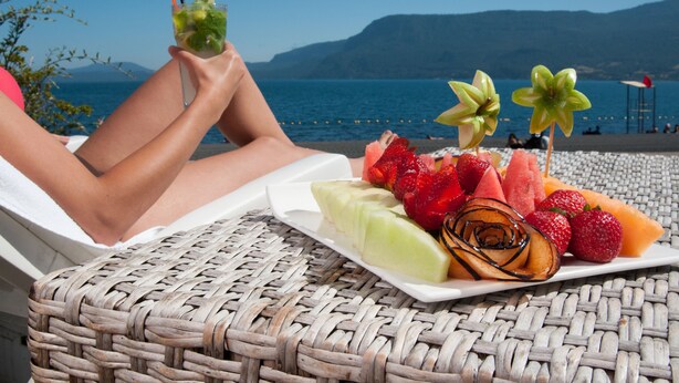 A wicker lounge chair with a plate of fruit and a drink, overlooking a beach with people in the background. - Enjoy Pucon (Pucón)