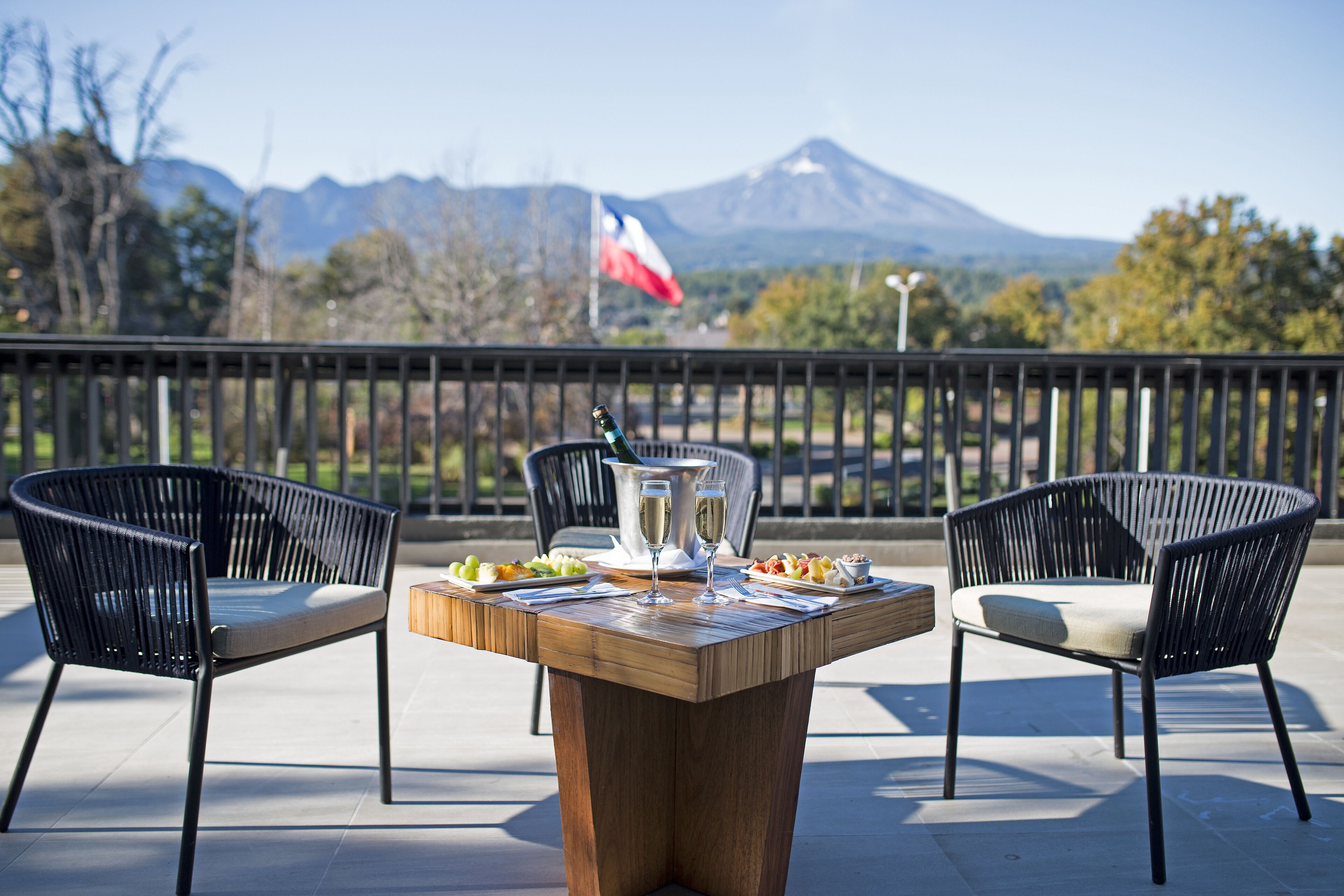 A wooden table with two black chairs, a bottle, and two glasses on it, set against a scenic mountain view with a flag in the background.