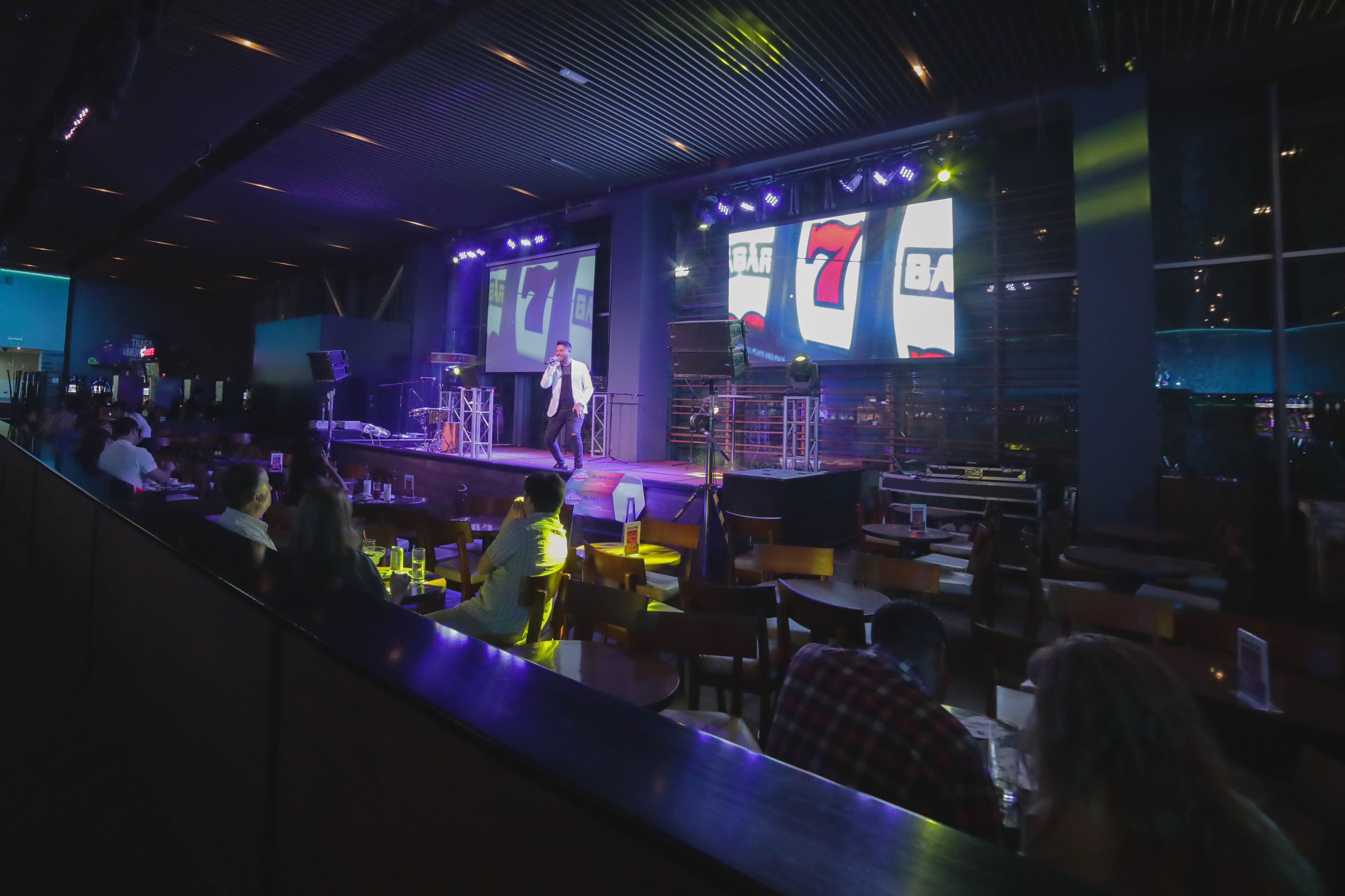Un escenario con un artista, una pantalla grande mostrando 'BAR 7', y una zona de bar con mesas y sillas.
