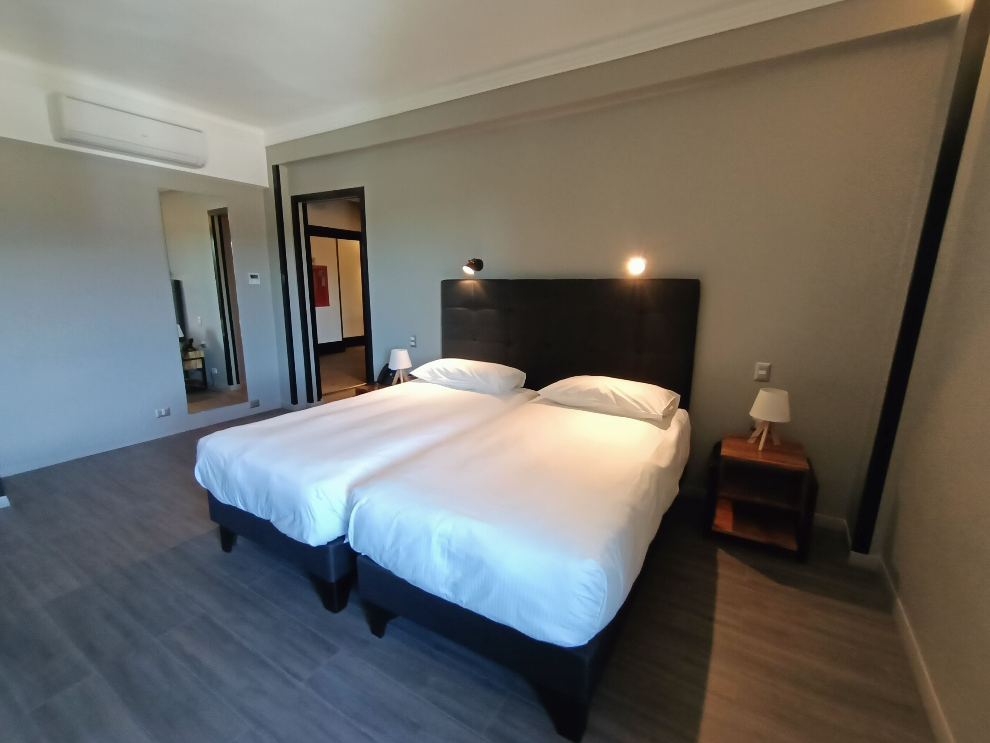 A modern hotel room with a large bed, bedside tables, a mirror, and an air conditioning unit.