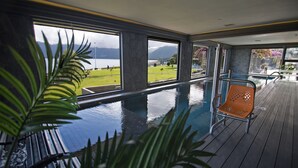 An indoor swimming pool with a view of a lake and mountains, surrounded by large windows and a wooden deck. - Enjoy Pucon (Pucón)