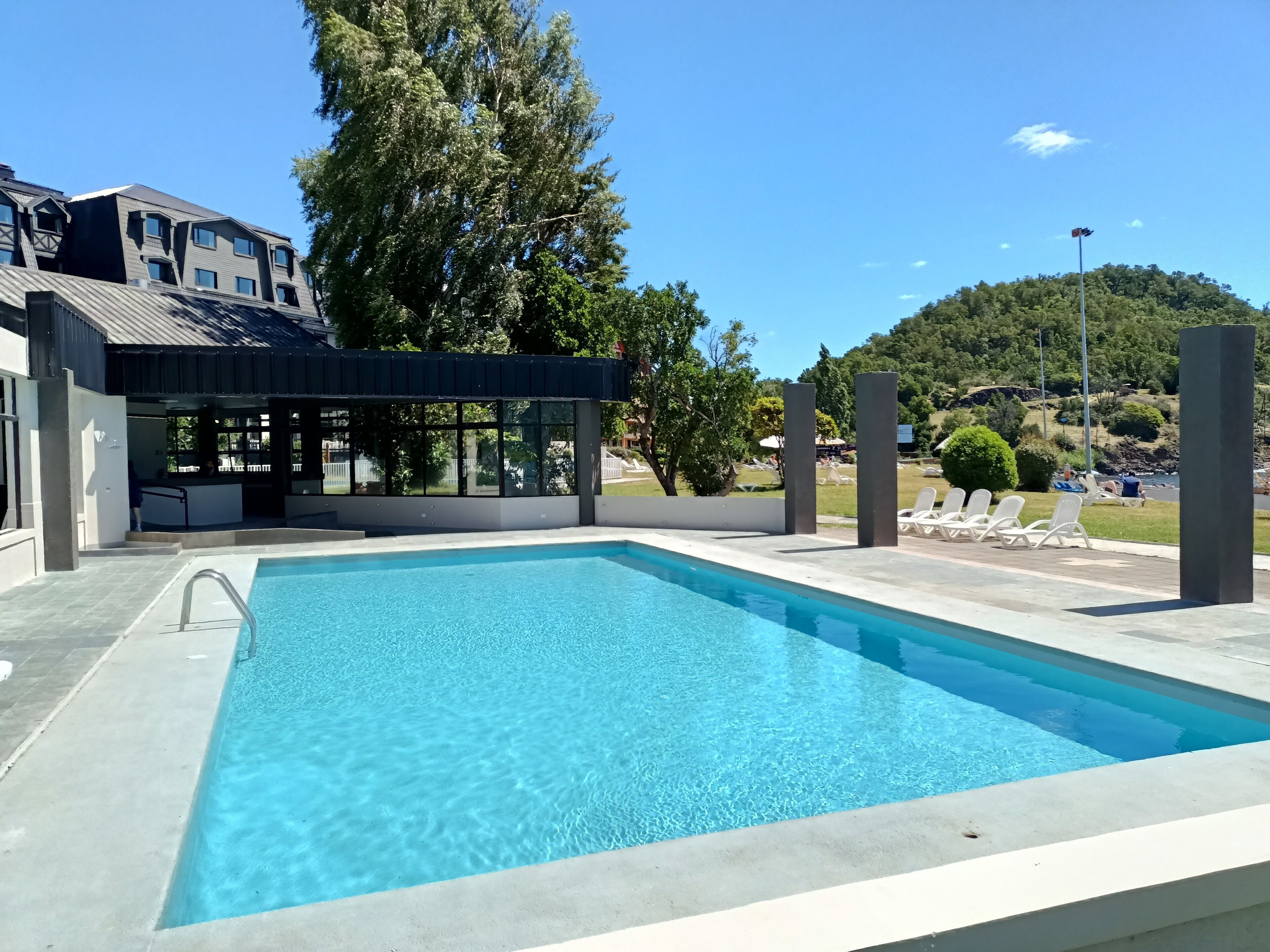 Une piscine rectangulaire à l’eau d’un bleu limpide, entourée d’une terrasse en béton. On y trouve des chaises longues blanches et un espace salon couvert avec des murs en verre.