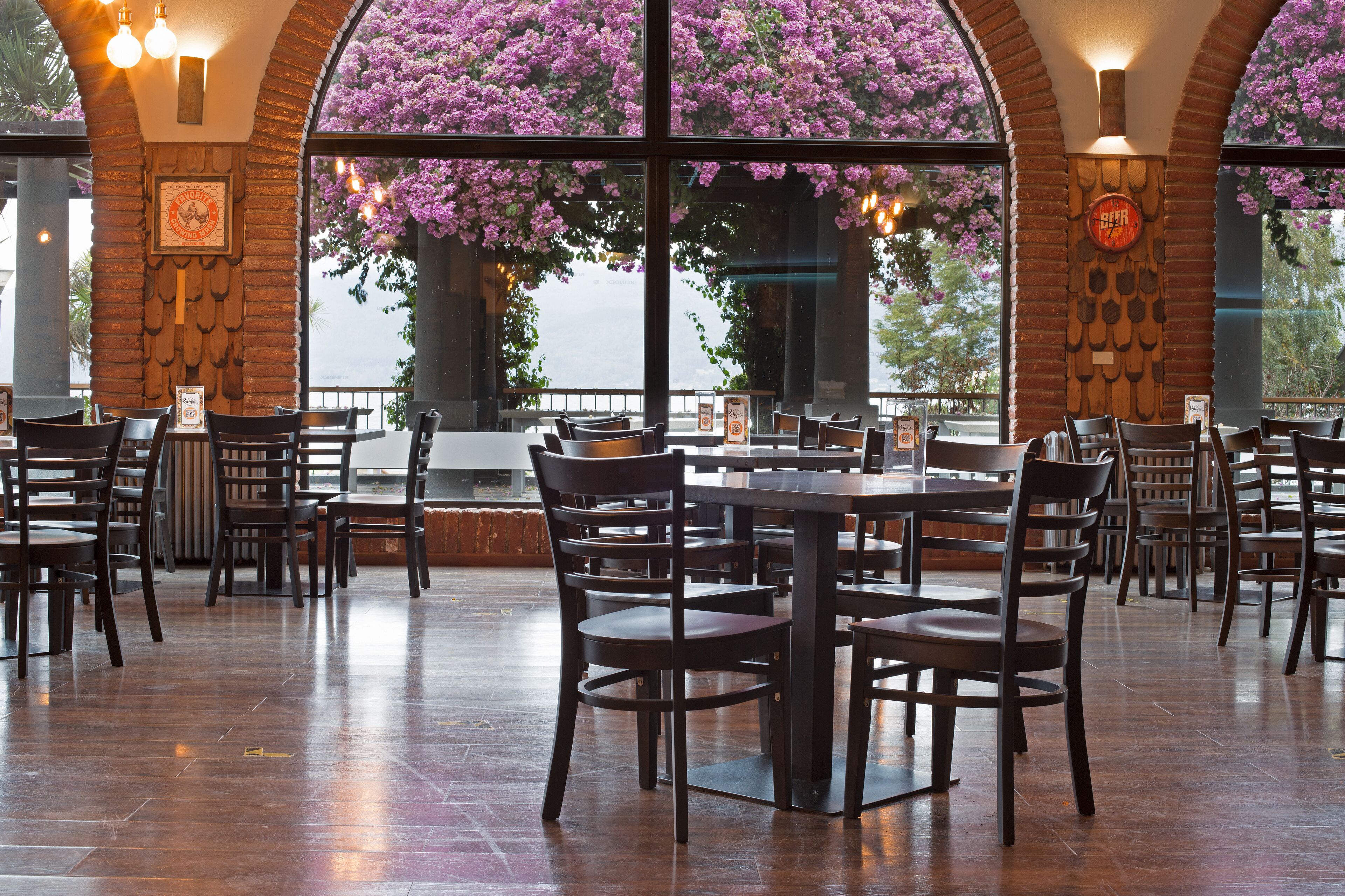 Een restaurant met bakstenen muren, grote ramen en houten tafels en stoelen.