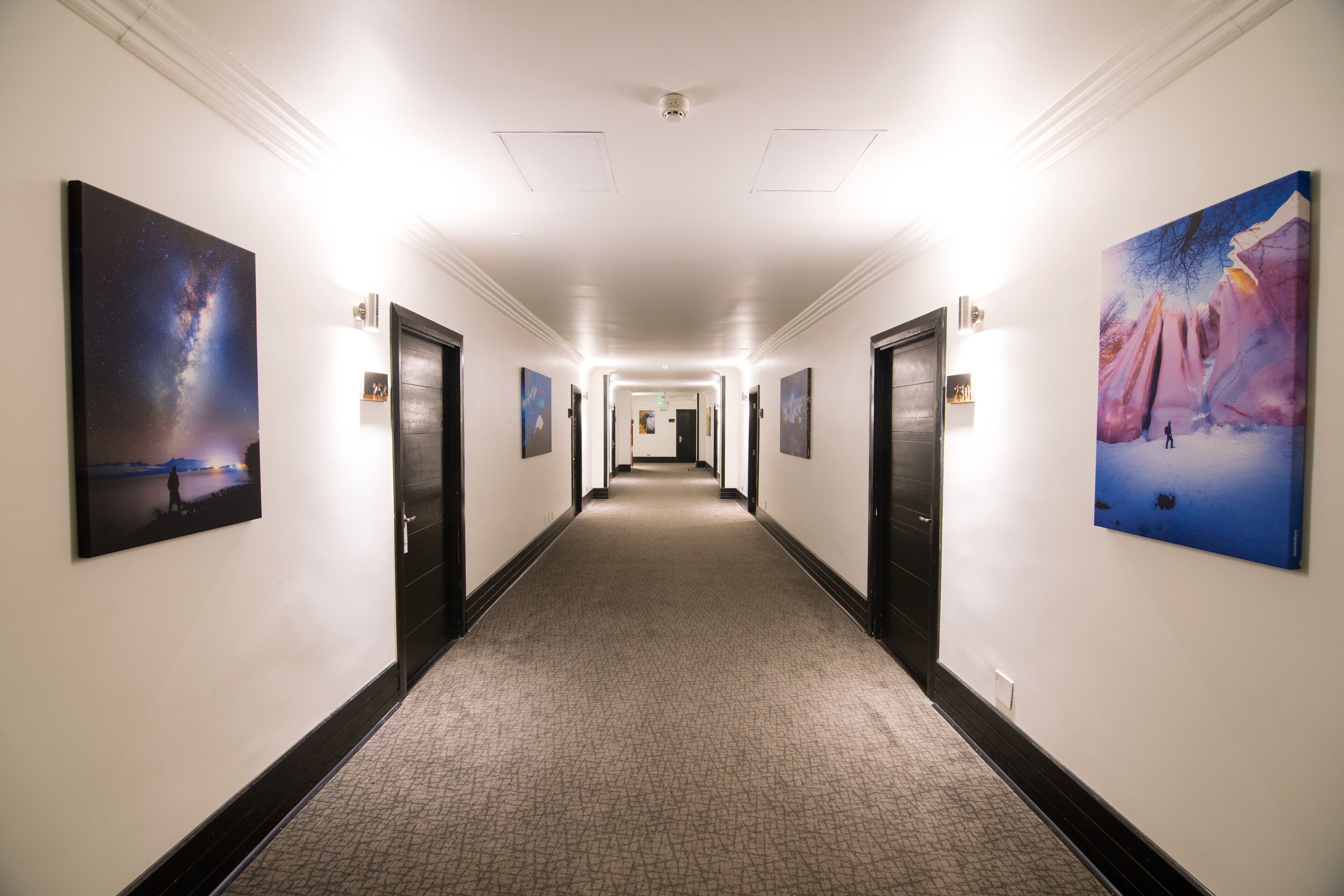 A hotel hallway with a carpeted floor, framed artwork on the walls, and doors leading to rooms.
