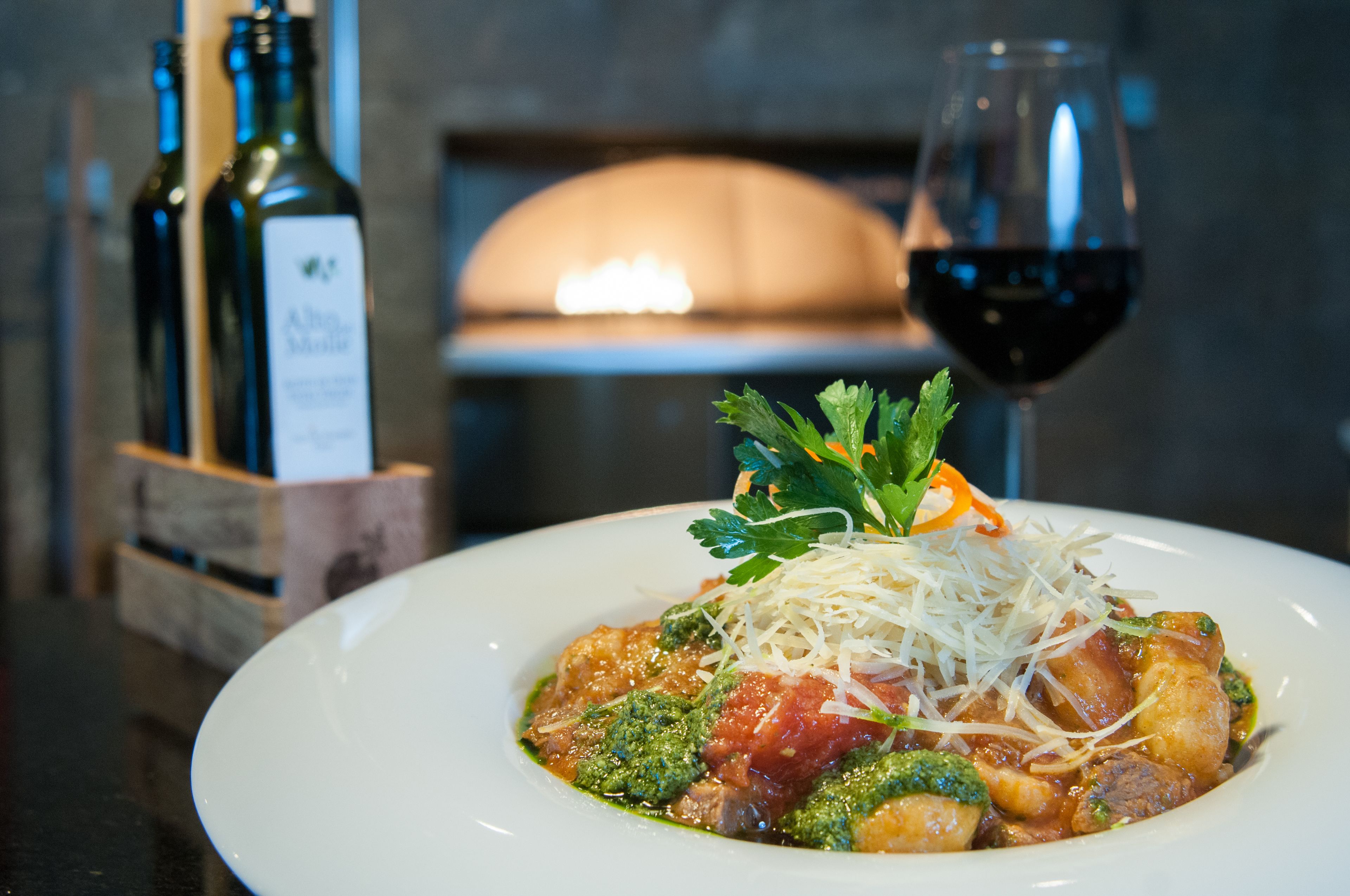 A plate of pasta with a garnish, a glass of red wine, and two bottles of wine in the background.