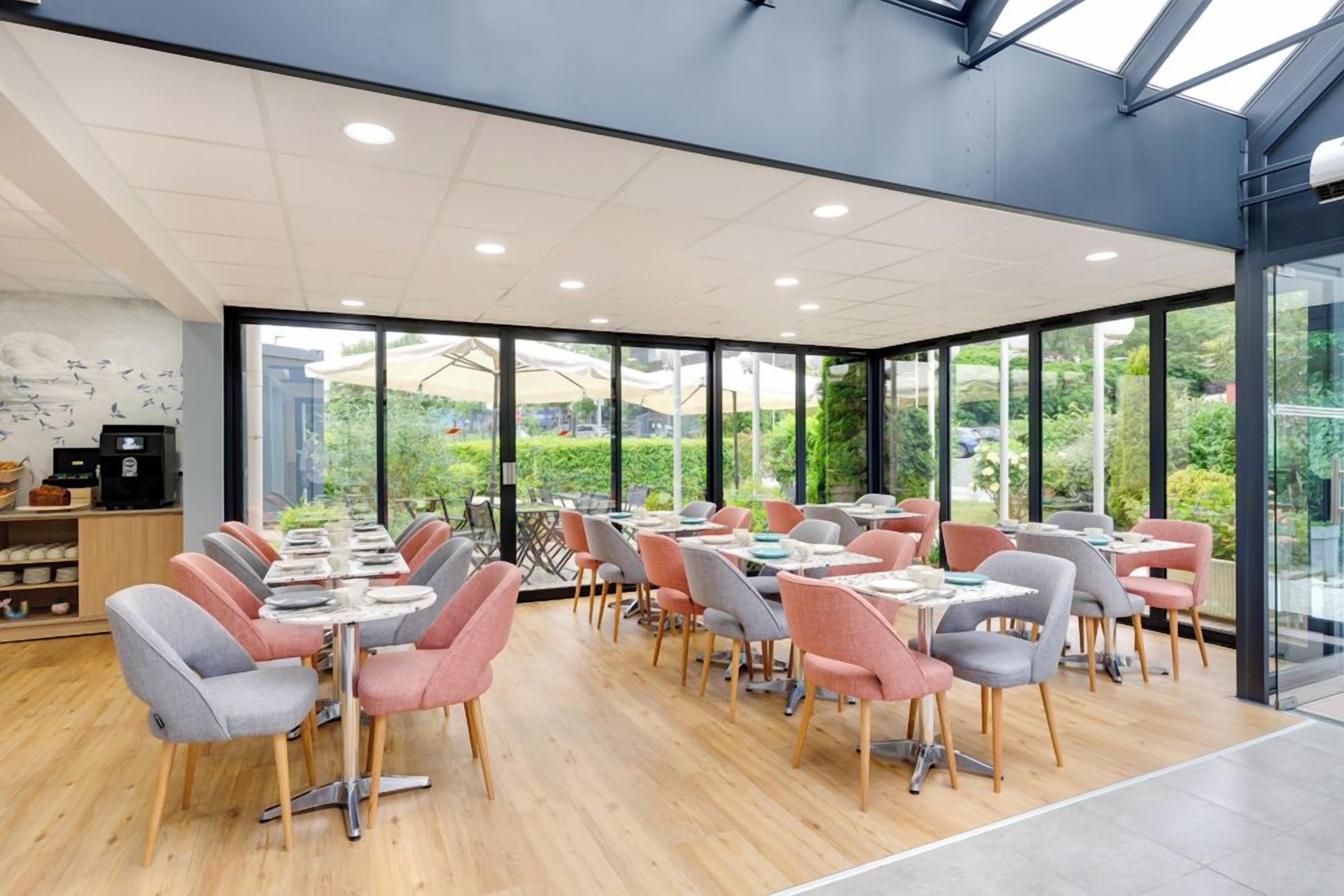 A modern dining area with tables and chairs, large windows, and a view of greenery outside.