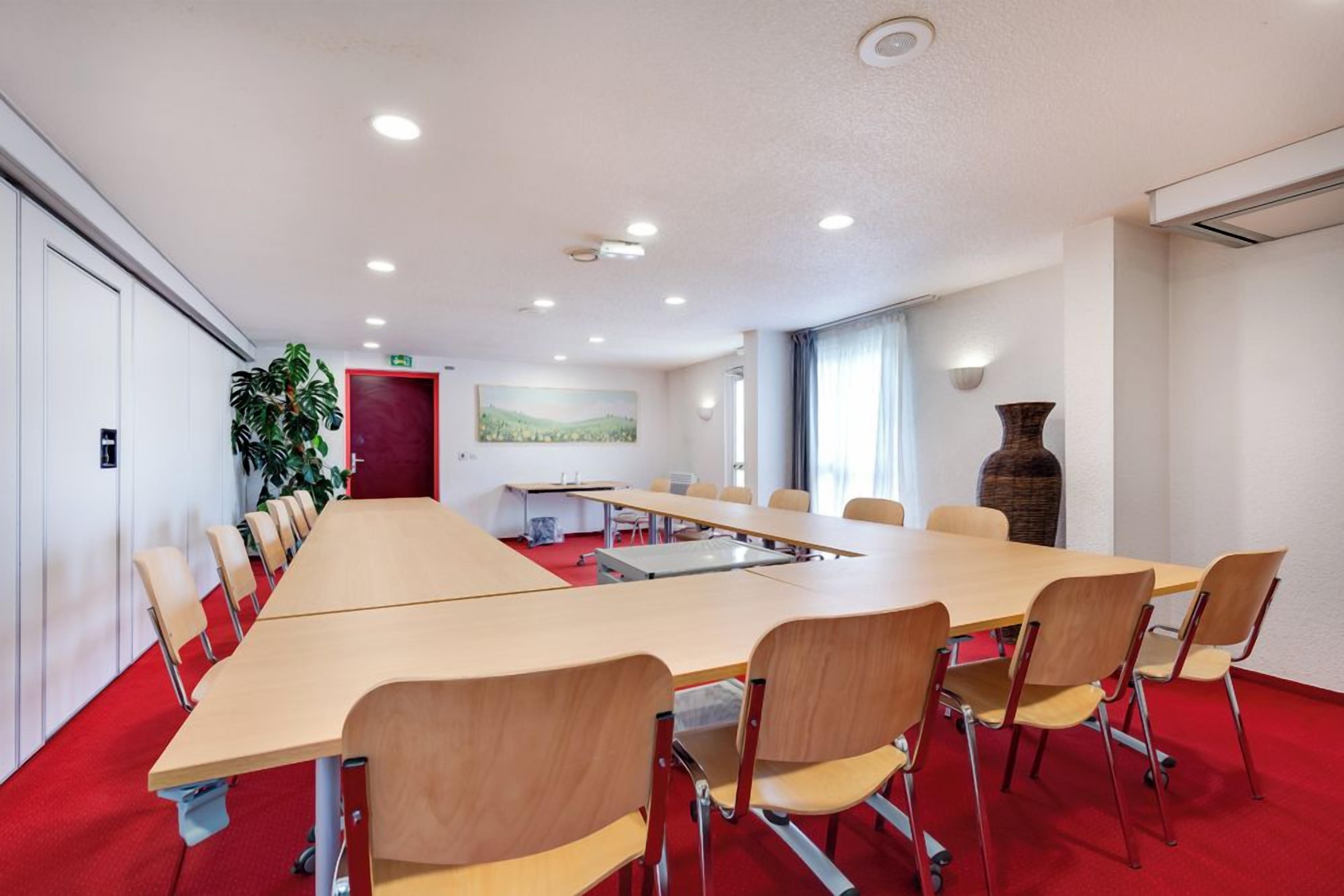 Une salle de conférence avec une longue table, des chaises, une moquette rouge, une plante, un tableau et un vase.