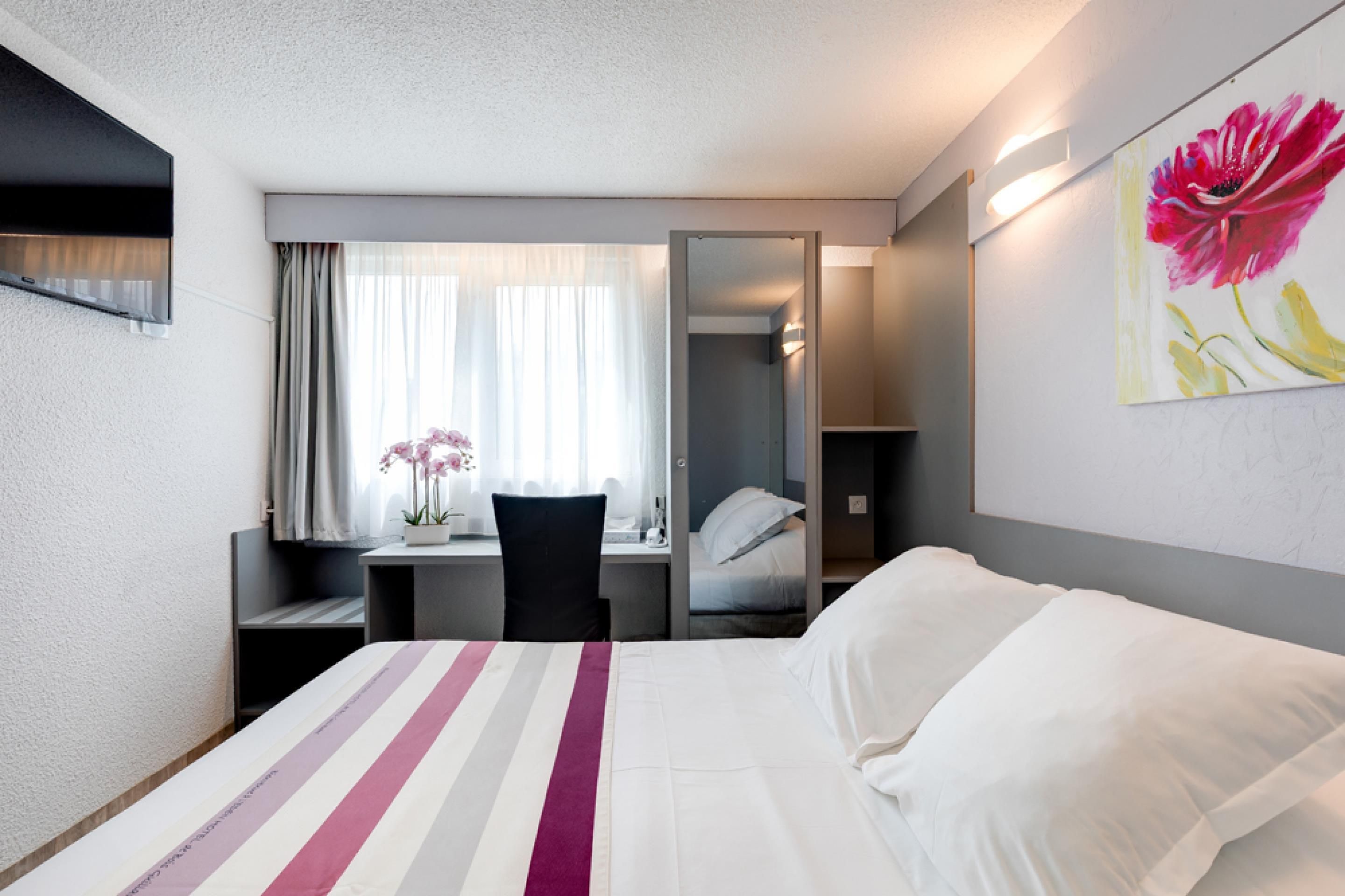 Une chambre d’hôtel avec un lit, un bureau, une chaise, une fenêtre avec des rideaux, un miroir et un tableau au mur.