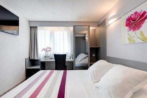A hotel room with a bed, a desk, a chair, a window with curtains, a mirror, and a painting on the wall. - Brit Hotel Confort Rouen Nord - Eden (Bois-Guillaume)