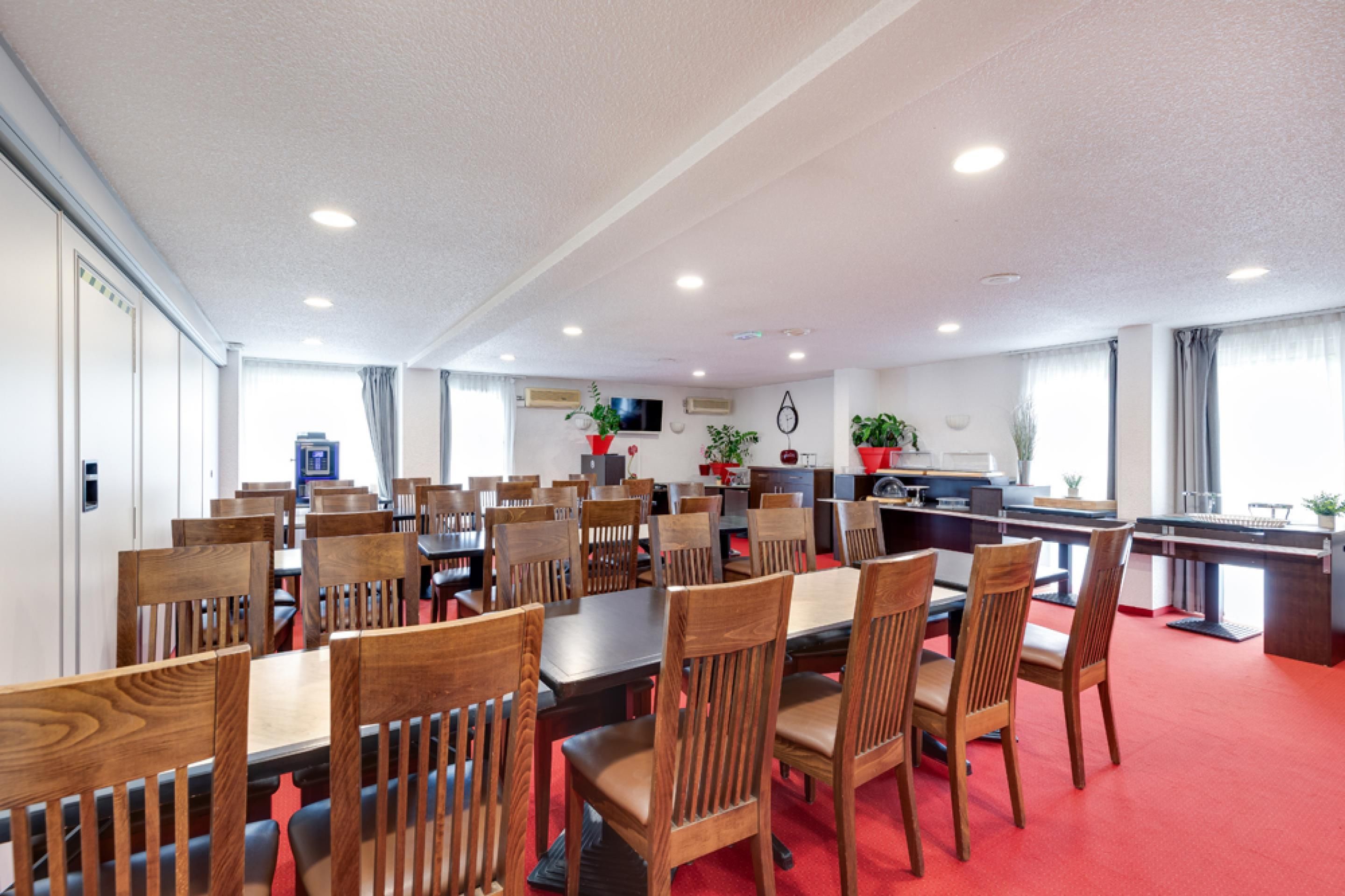 A hotel dining area with multiple wooden tables and chairs, a kitchen area, and a red carpeted floor.