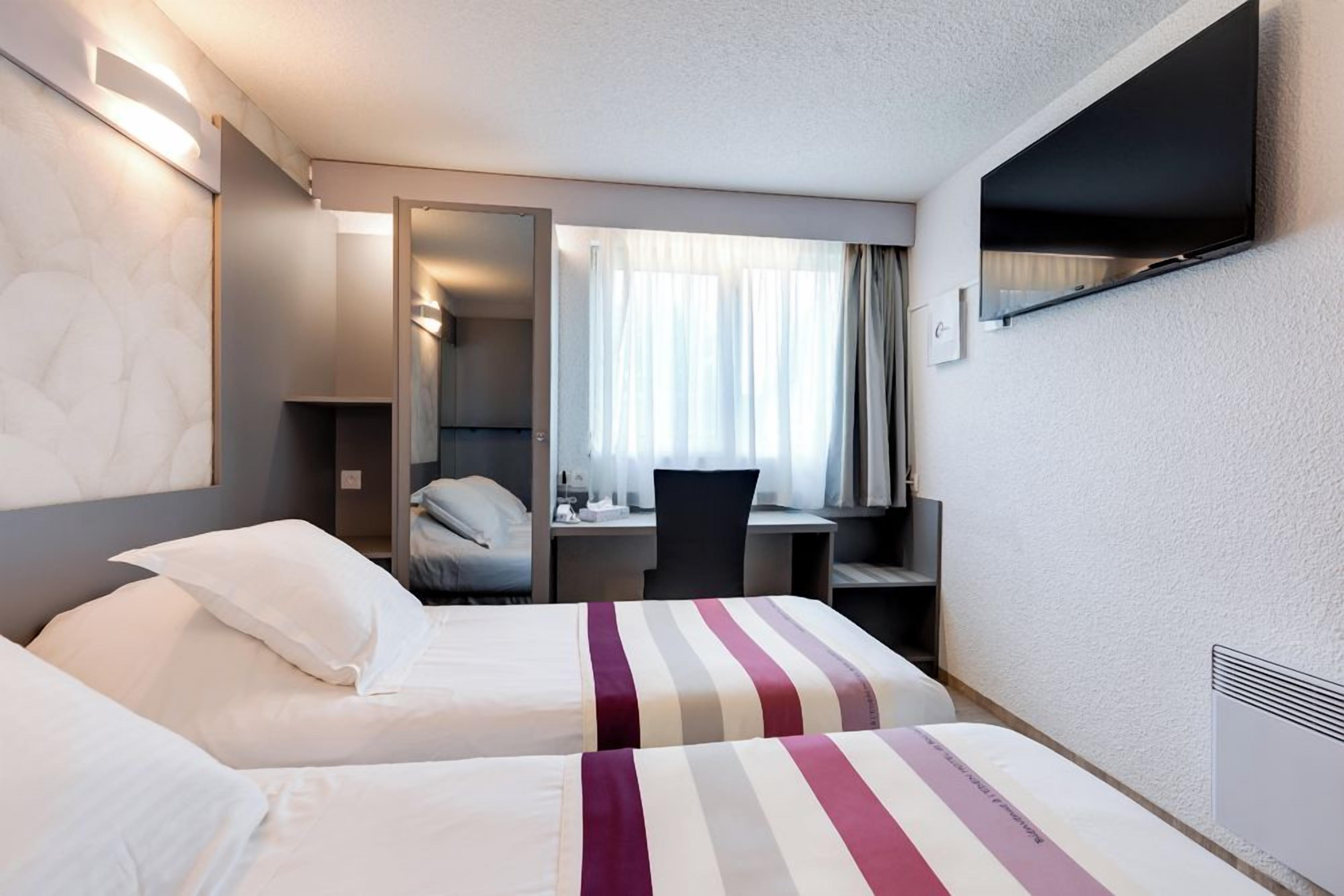 Quarto de hotel com duas camas, uma escrivaninha, uma cadeira, televisão fixada na parede e janela com cortinas.