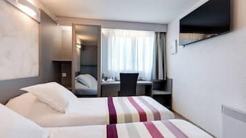 Quarto de hotel com duas camas, uma escrivaninha, uma cadeira, televisão fixada na parede e janela com cortinas.