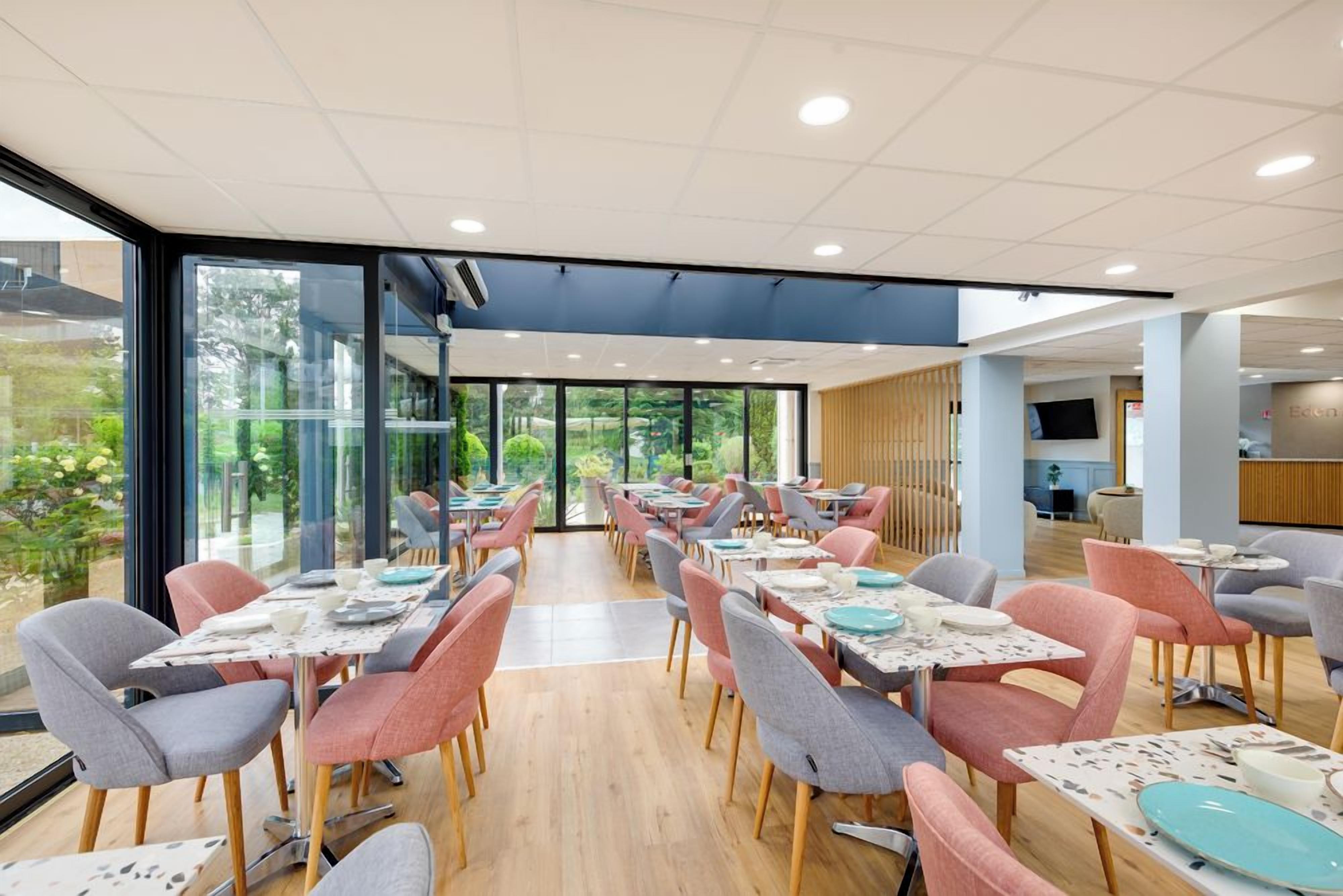 Een moderne eetruimte met tafels en stoelen, grote ramen en uitzicht op groen.