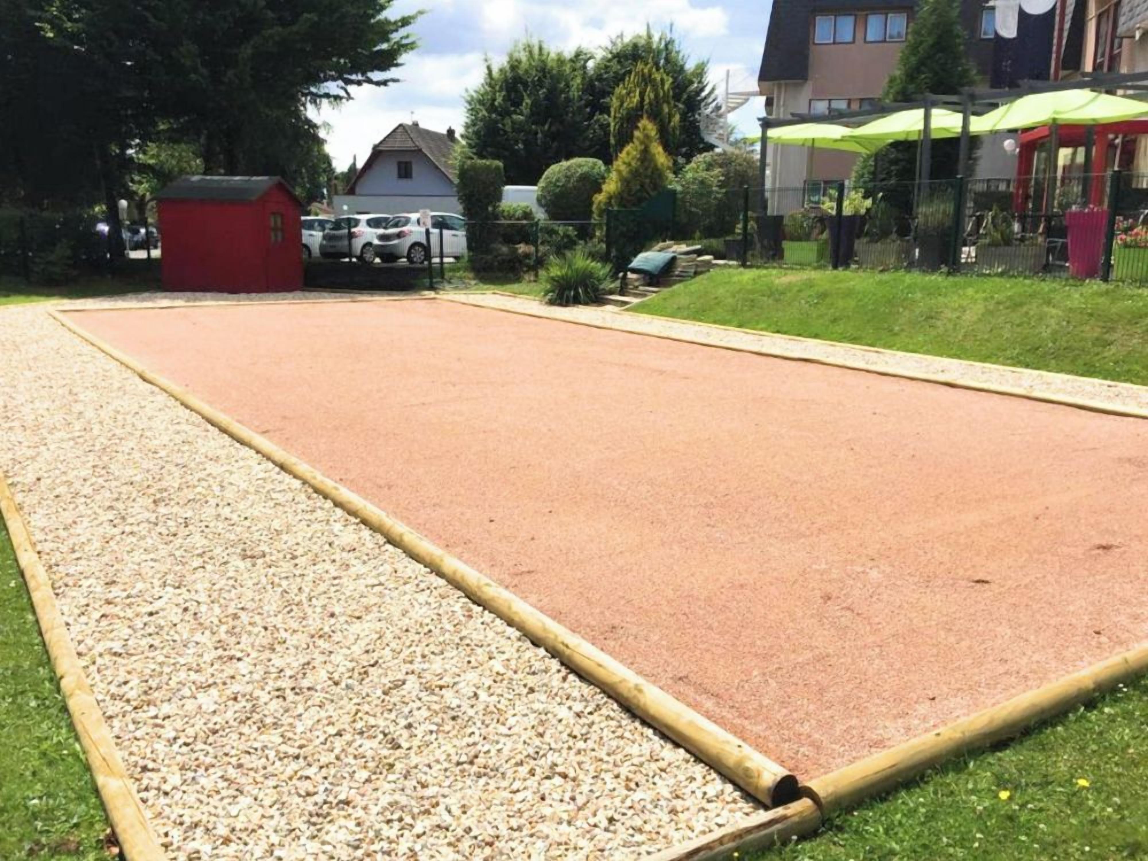 Ein großer Außenbereich mit Kiesweg und roter Fläche, möglicherweise ein Sportplatz, umgeben von Grünflächen und Gebäuden.