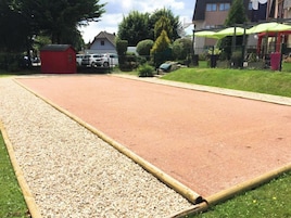 Ampla área externa com um caminho de cascalho e superfície vermelha, possivelmente uma quadra esportiva, cercada por vegetação e edifícios.