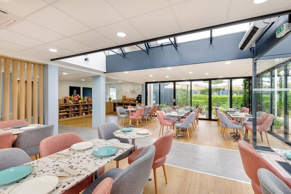 A spacious dining area with multiple tables and chairs, a children's play area in the background, and large windows providing a view of the outdoors.