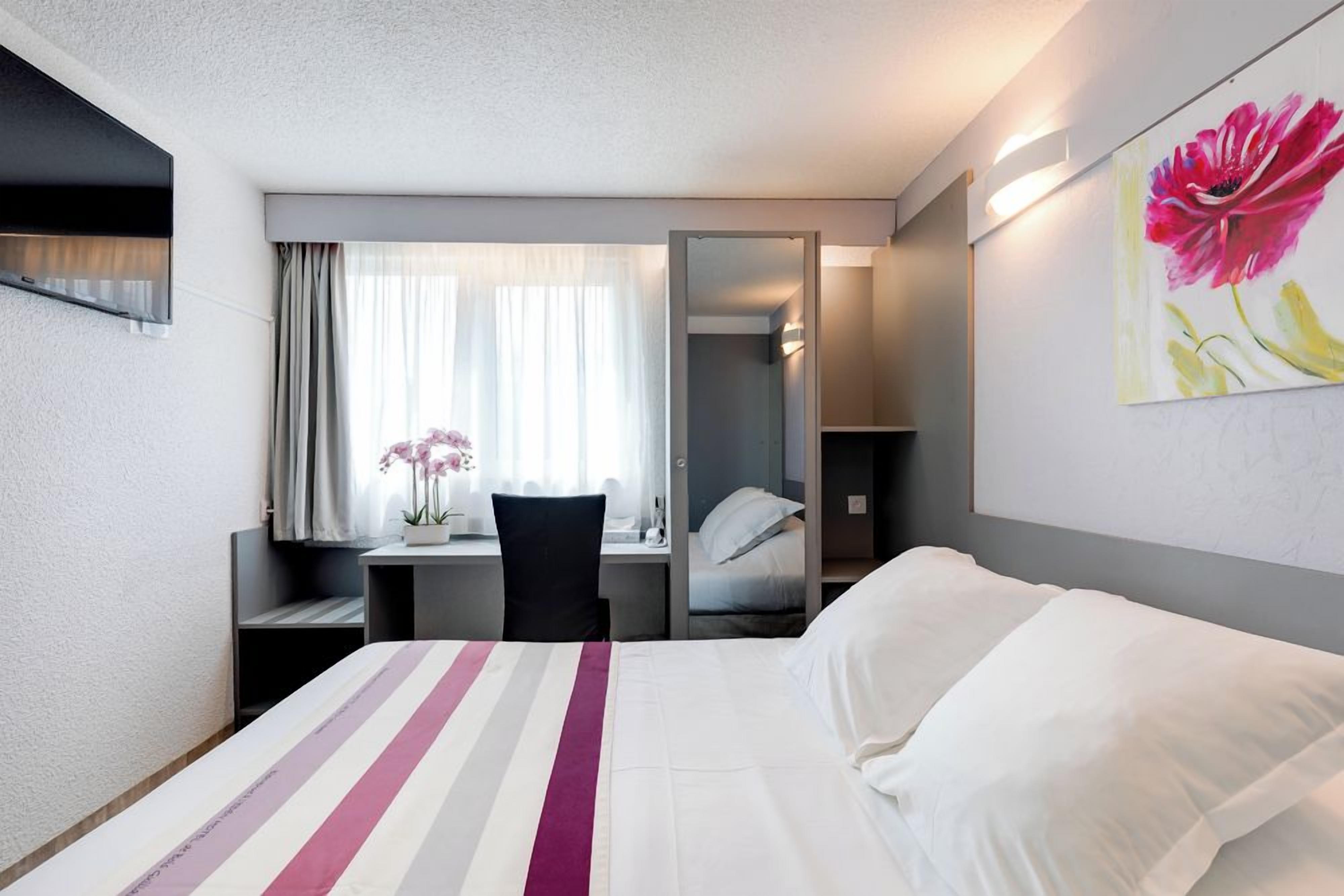 Ett hotellrum med en säng, ett skrivbord med en stol, ett fönster med gardiner, en spegel och en tavla på väggen.