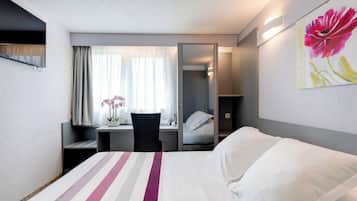 A hotel room with a bed, a desk with a chair, a window with curtains, a mirror, and a painting on the wall.