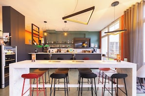 Un bar moderno con mostrador de mármol, varios taburetes, lámparas colgantes y un amplio ventanal que proporciona luz natural.