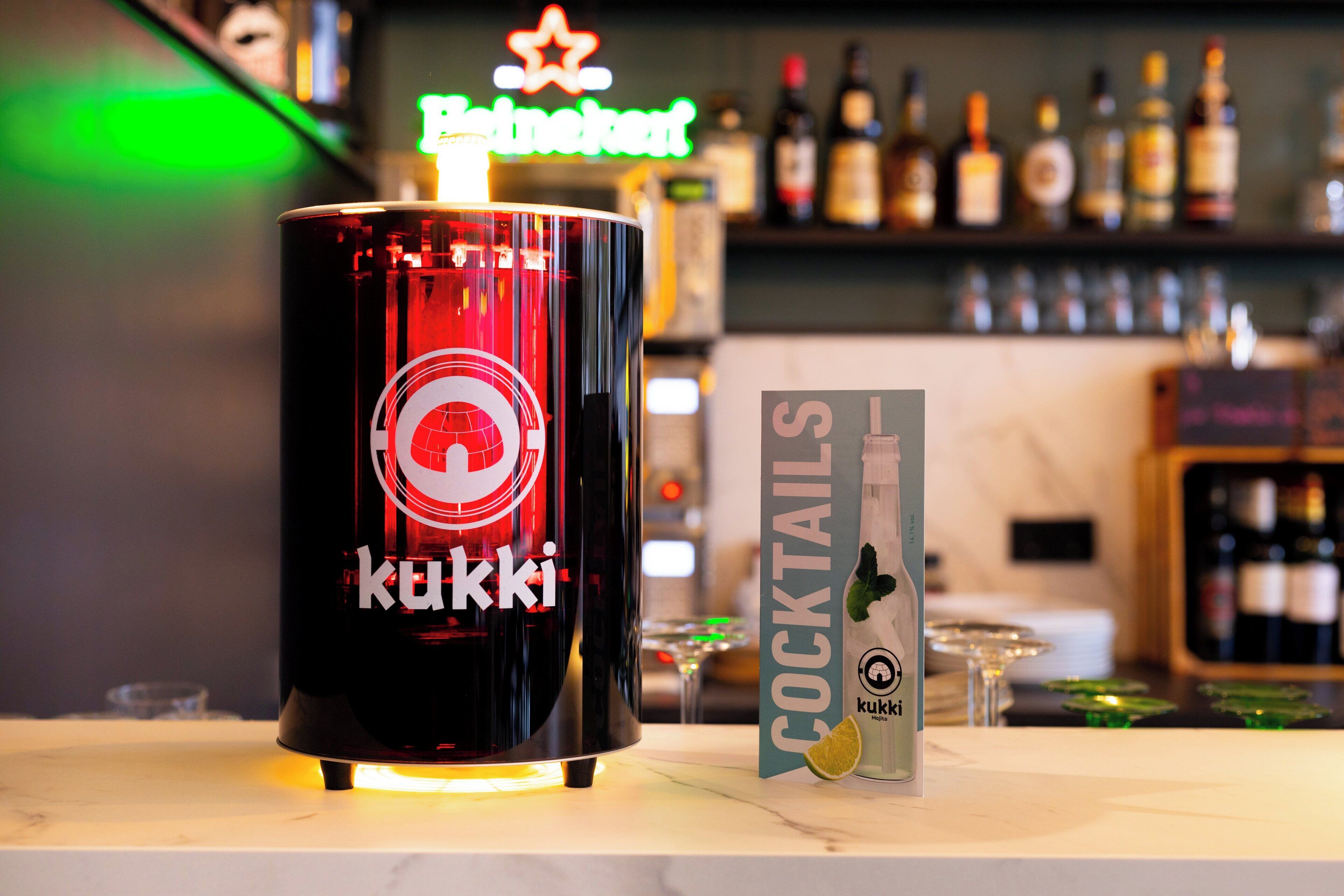 A bar area with a KUKKI branded cocktail shaker, a sign for COCKTAILS, and a neon sign for Heineken in the background.