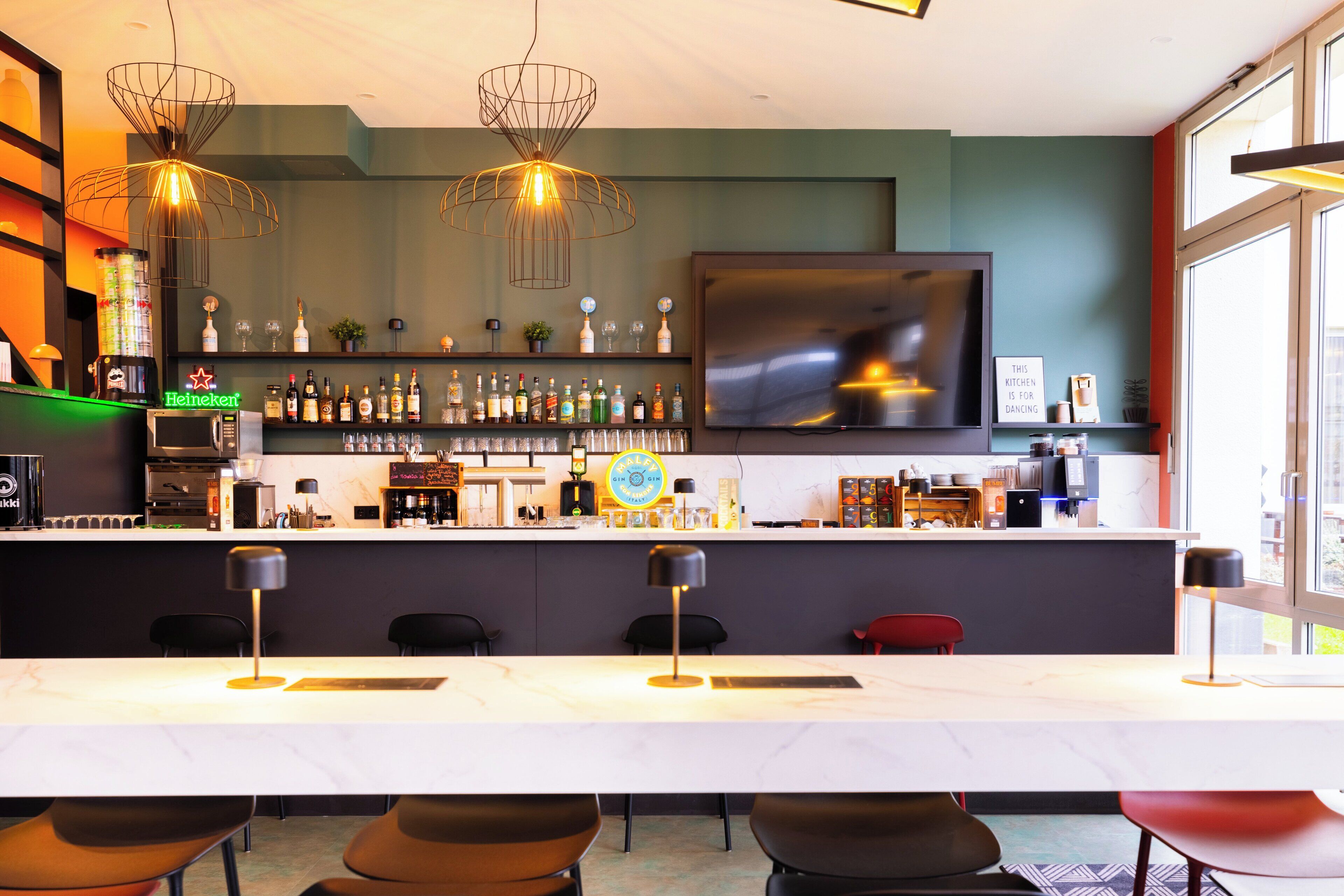 Un área de bar moderna con una encimera blanca, varias botellas de licor, un letrero de Heineken y un televisor montado en la pared.