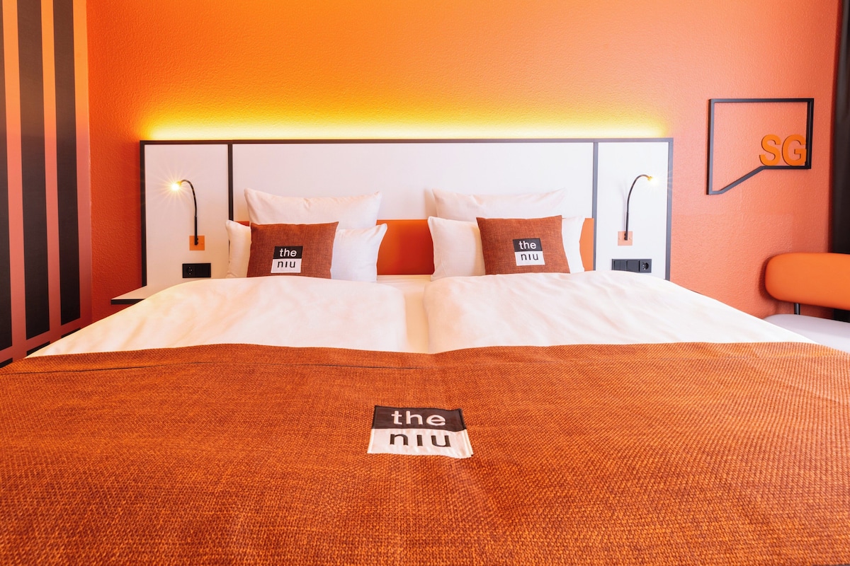 Habitación de hotel con una cama grande, paredes anaranjadas y cabecera con un logotipo.