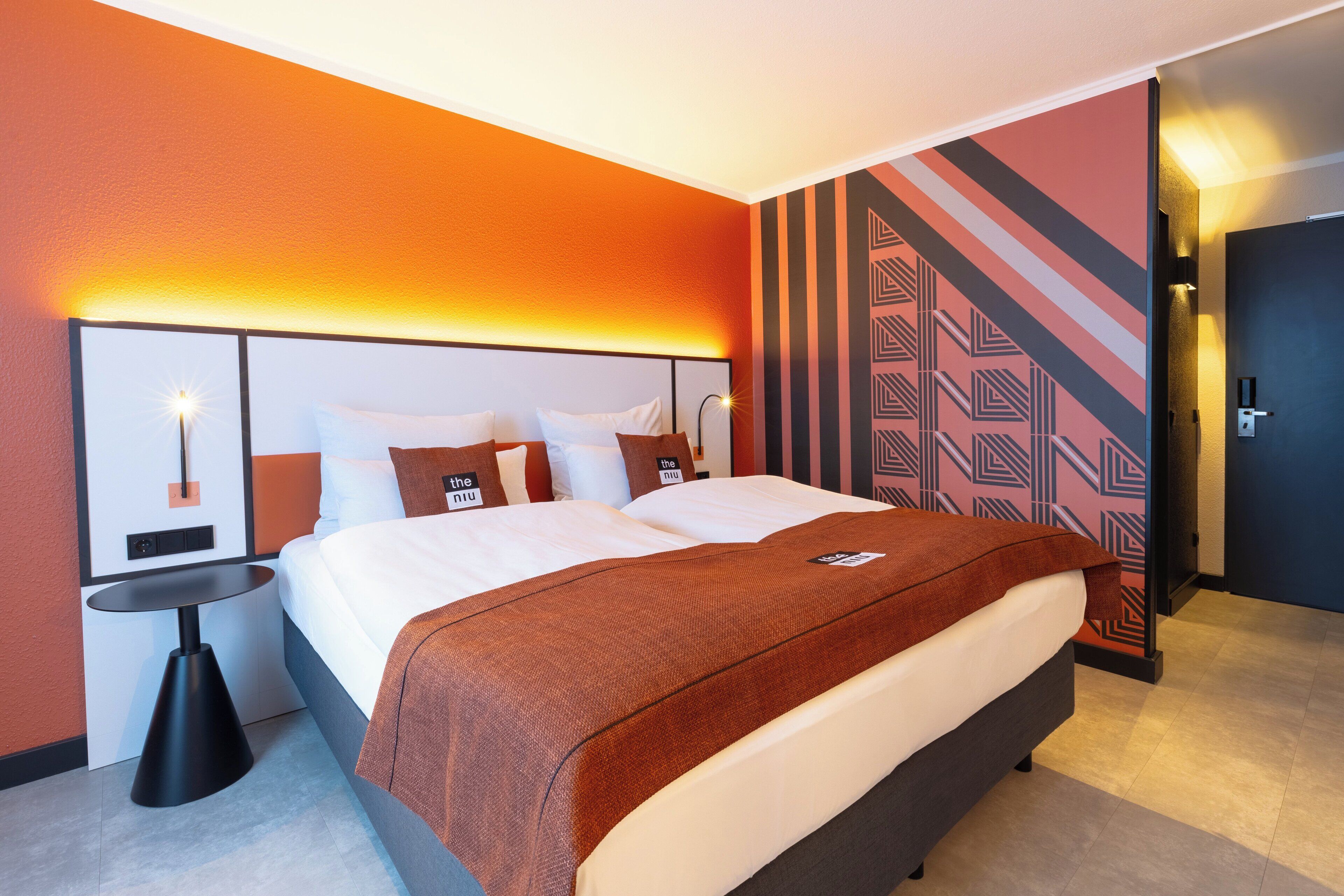 A modern hotel room with a large bed, a bedside table, and a wall with geometric patterns.