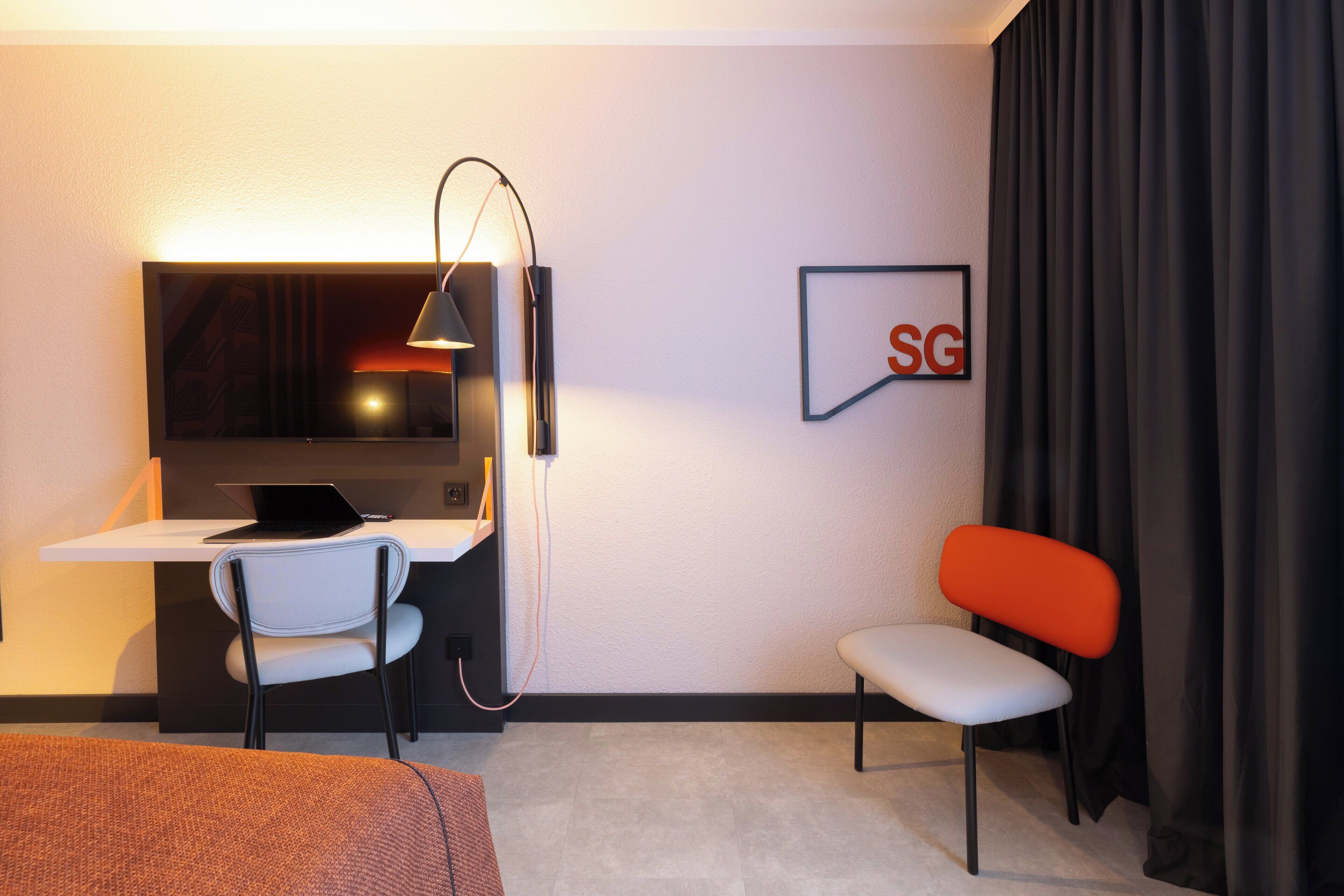 A hotel room with a desk, a laptop, a chair, a bed, and a framed picture on the wall.