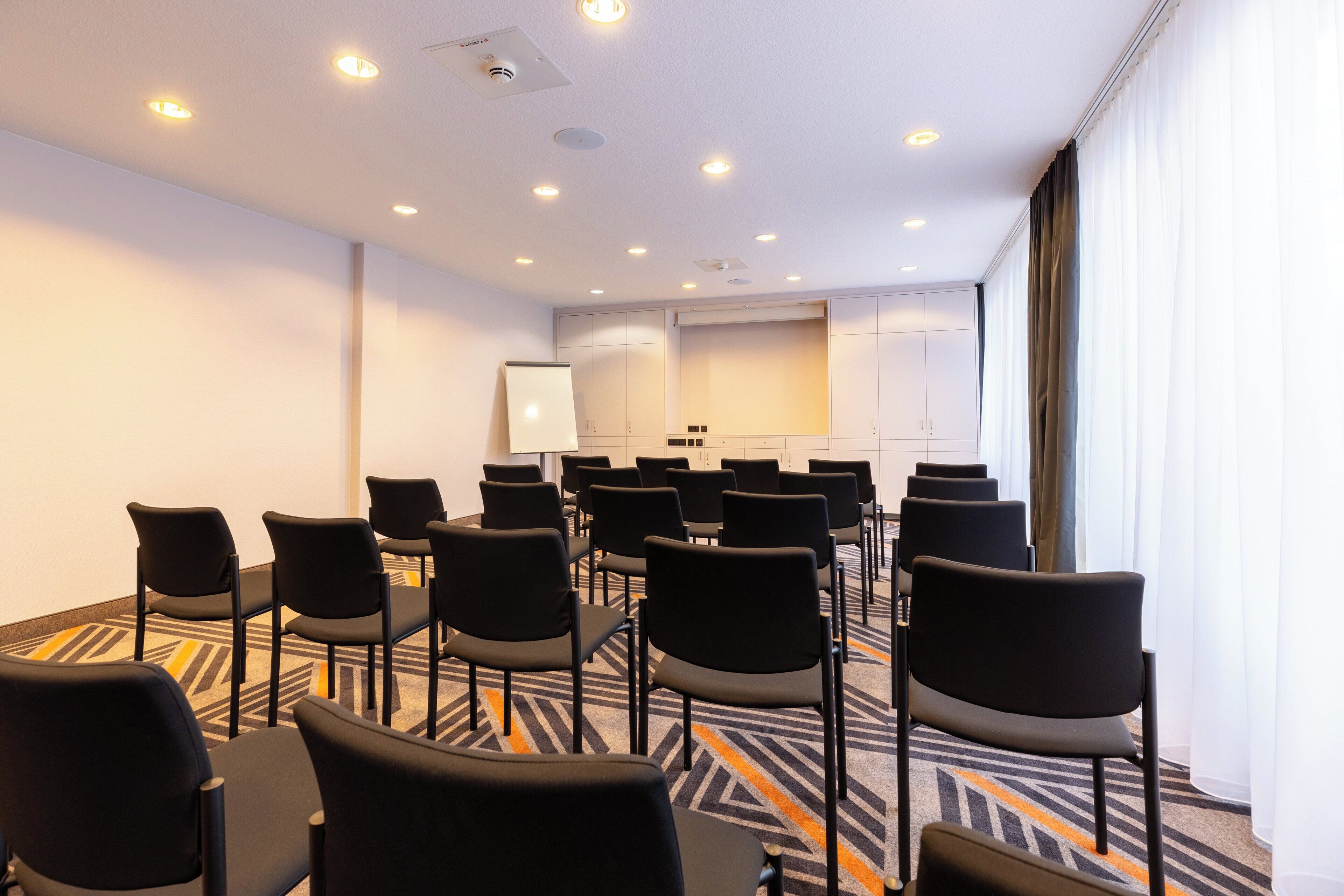 Uma sala de conferências com fileiras de cadeiras pretas, um quadro branco e um amplo janelão com cortinas.