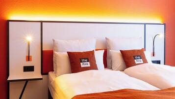 A hotel room with a bed, two pillows with text, a nightstand with a lamp, and a wall-mounted light.