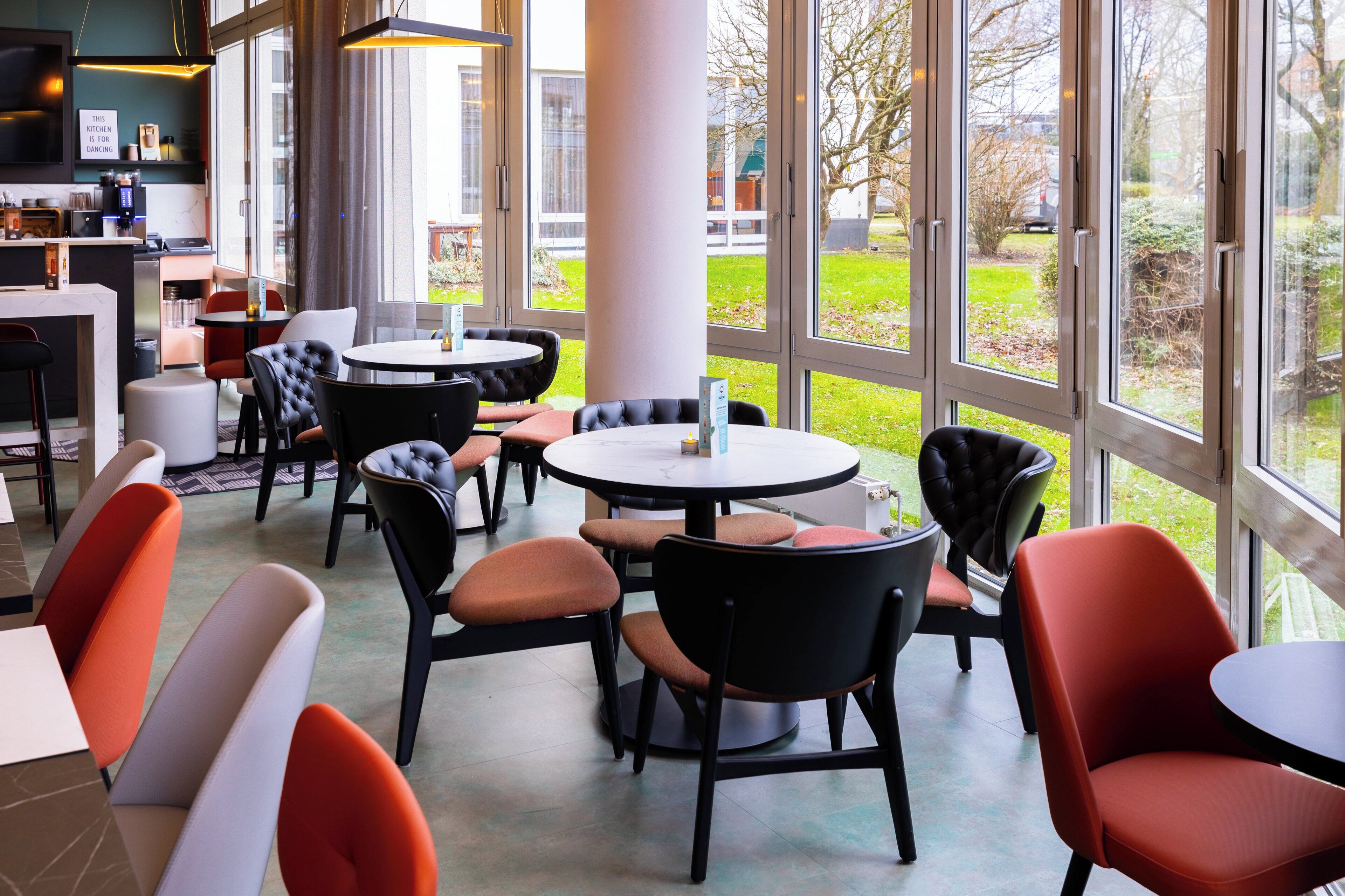 A modern restaurant with round tables and chairs, large windows offering a view of the outdoors, and a kitchen area visible in the background.