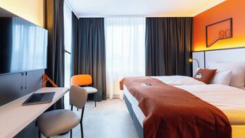 A hotel room with a bed, a desk with a laptop, a chair, a TV, and a window with curtains.