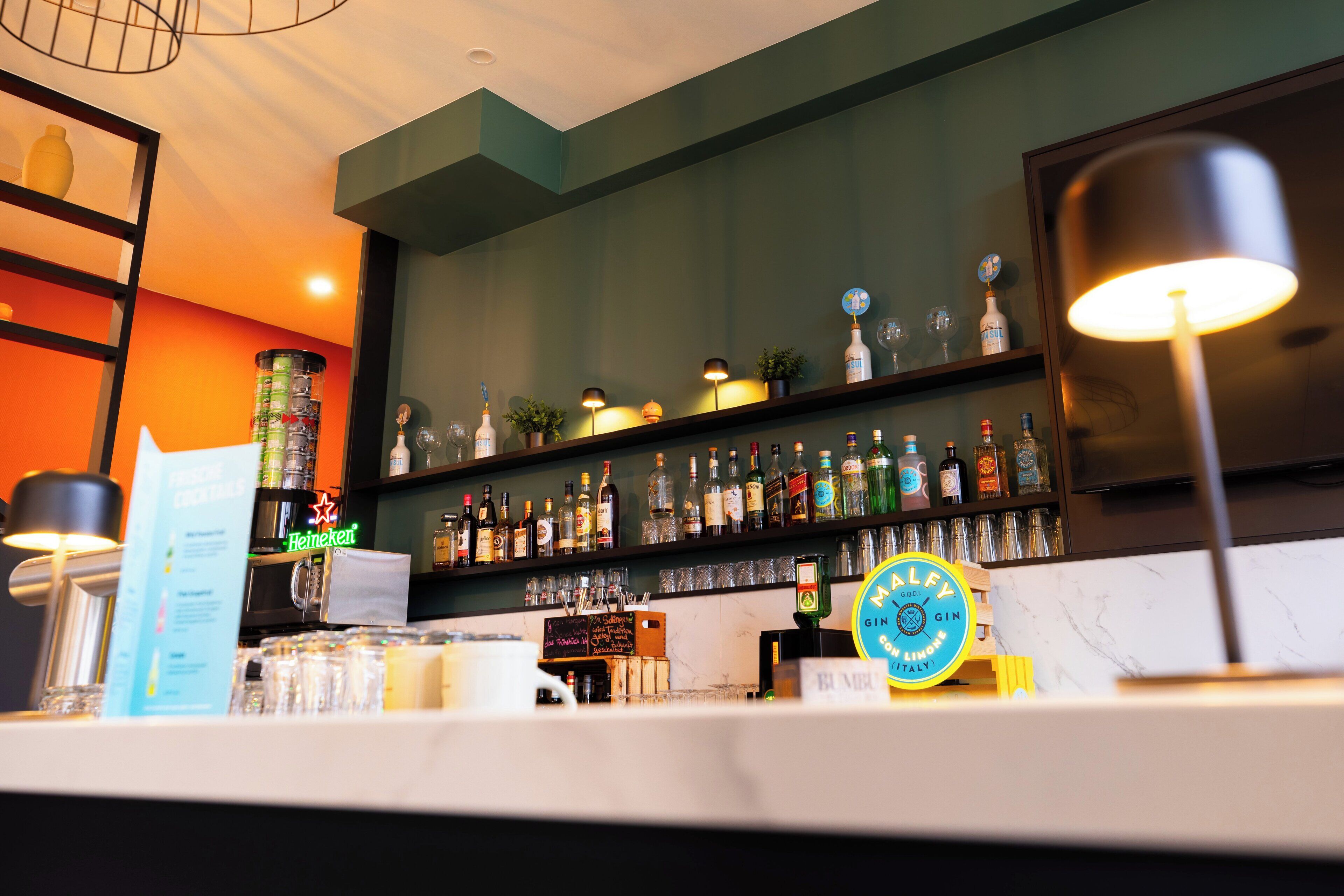 Un espace bar avec des étagères remplies de bouteilles de différentes boissons alcoolisées, une enseigne Heineken et un comptoir avec des verres et un menu.