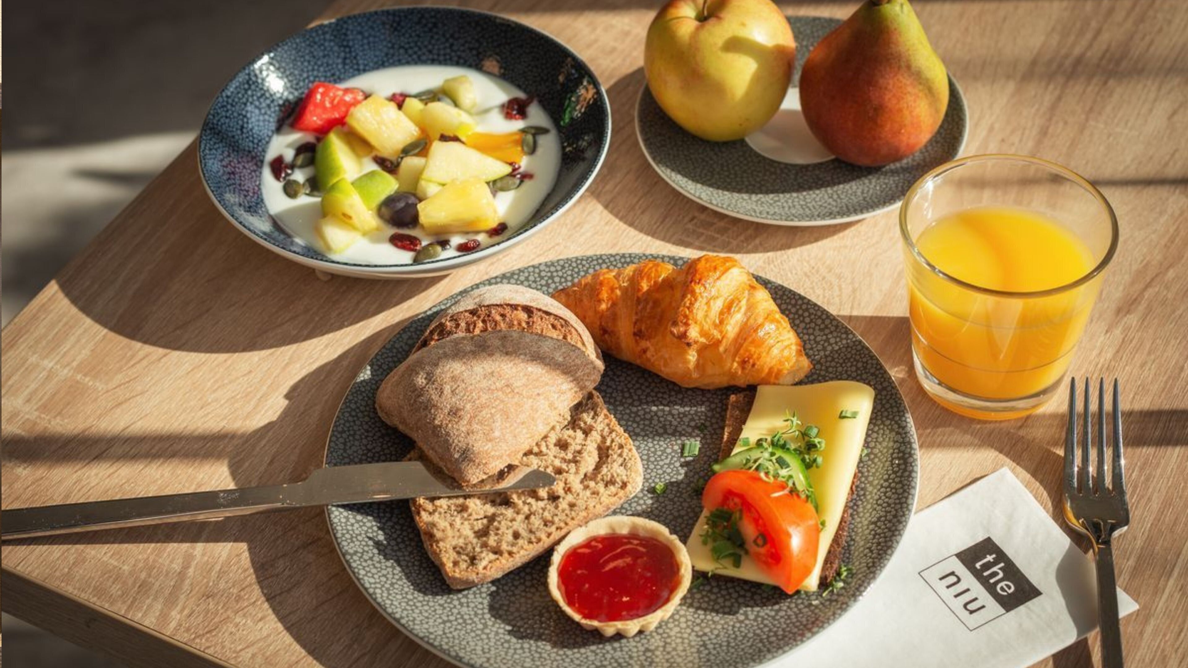 Un desayuno con un tazón de yogur y fruta, un croissant, una rebanada de pan con mantequilla, un sándwich de tomate y un vaso de zumo de naranja sobre una mesa de madera.