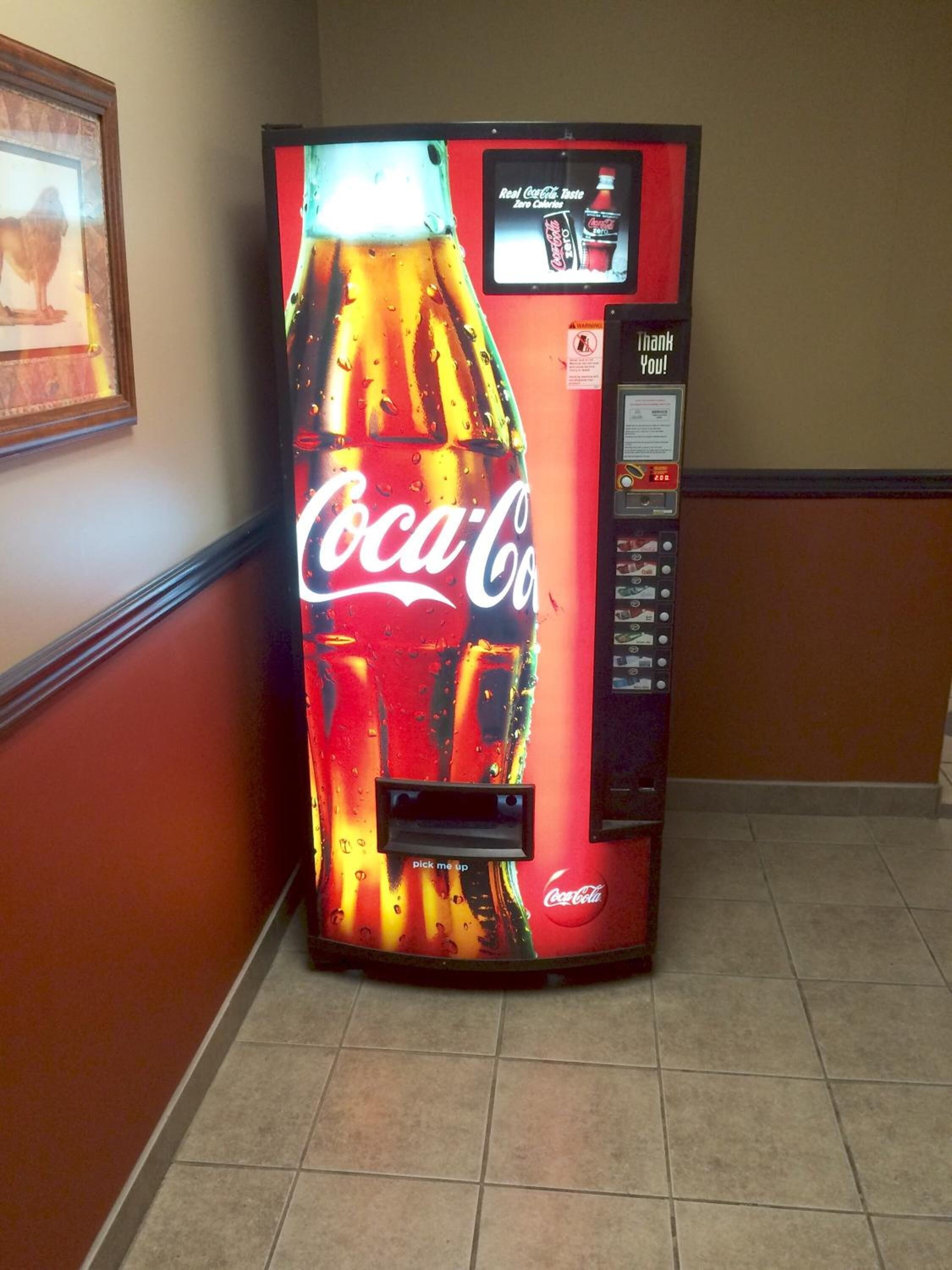 A Coca-Cola vending machine in a hallway with a framed picture on the wall.